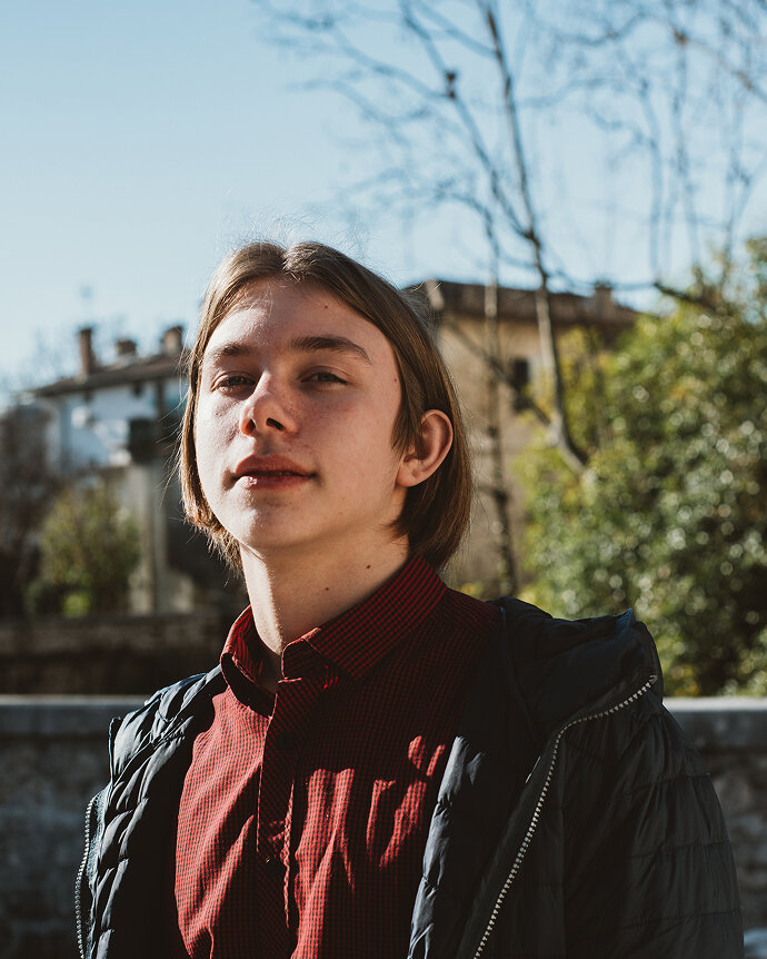 Junger Mensch in rot-kariertem Hemd und schwarzer Jacke vor einem verschwommenen Hintergrund mit B&auml;umen und Geb&auml;uden, in warmem Seitenlicht fotografiert.