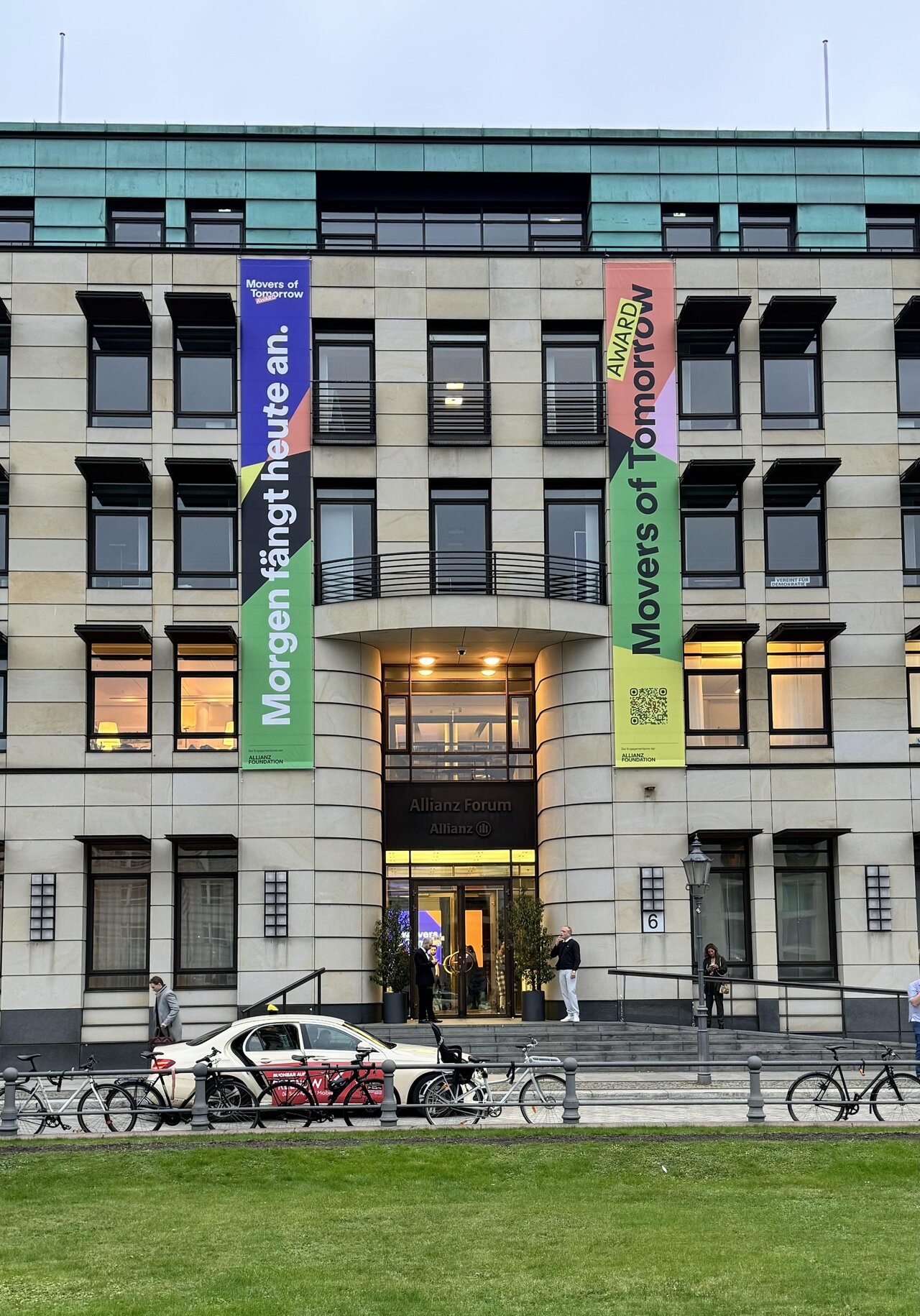 Moderne Fassade des Allianz Foundation-Geb&auml;udes mit bunten Bannern f&uuml;r den "Movers of Tomorrow Award" und Fahrr&auml;dern im Vordergrund, die den Eingang und die urbane Umgebung in Berlin zeigen.