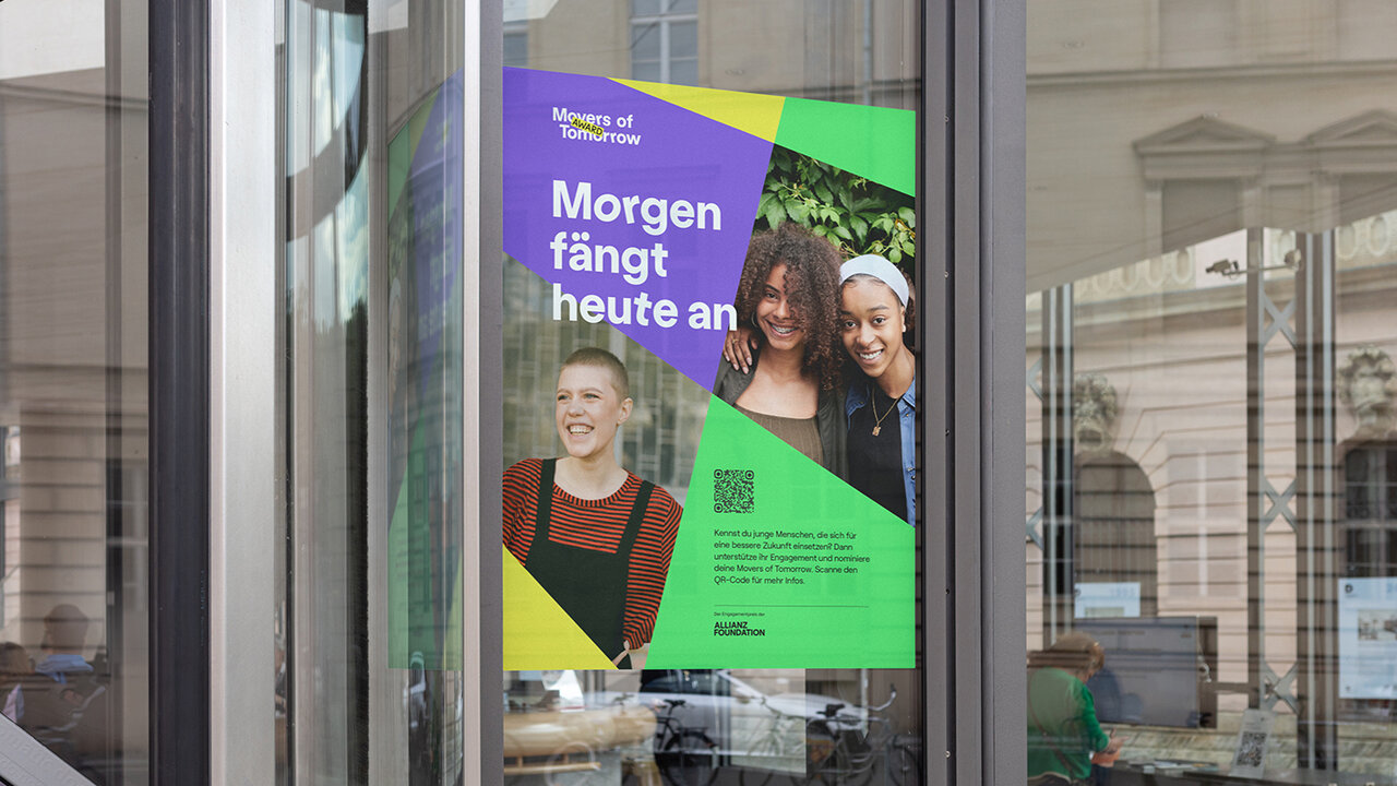 Werbeplakat f&uuml;r den "Movers of Tomorrow Award" mit drei l&auml;chelnden jungen Menschen auf gr&uuml;n-violettem Hintergrund, pr&auml;sentiert in einem Schaufenster mit Spiegelung einer Stadtarchitektur.