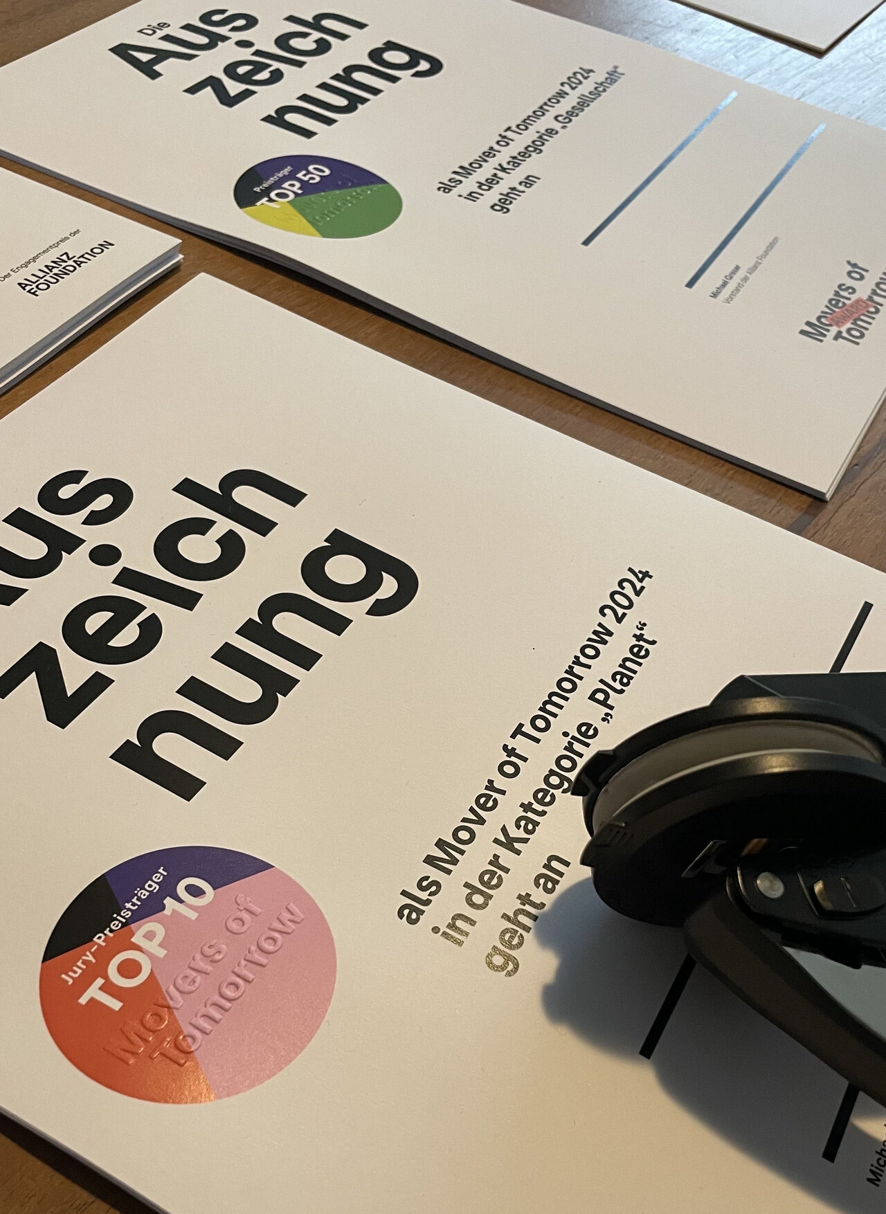 Zwei Brosch&uuml;ren des Allianz Foundation "Movers of Tomorrow Award" 2024 mit grafischem Farbdesign und einem Teil einer Kopfh&ouml;rer oder Kamera im Vordergrund, auf einem Holztisch liegend.