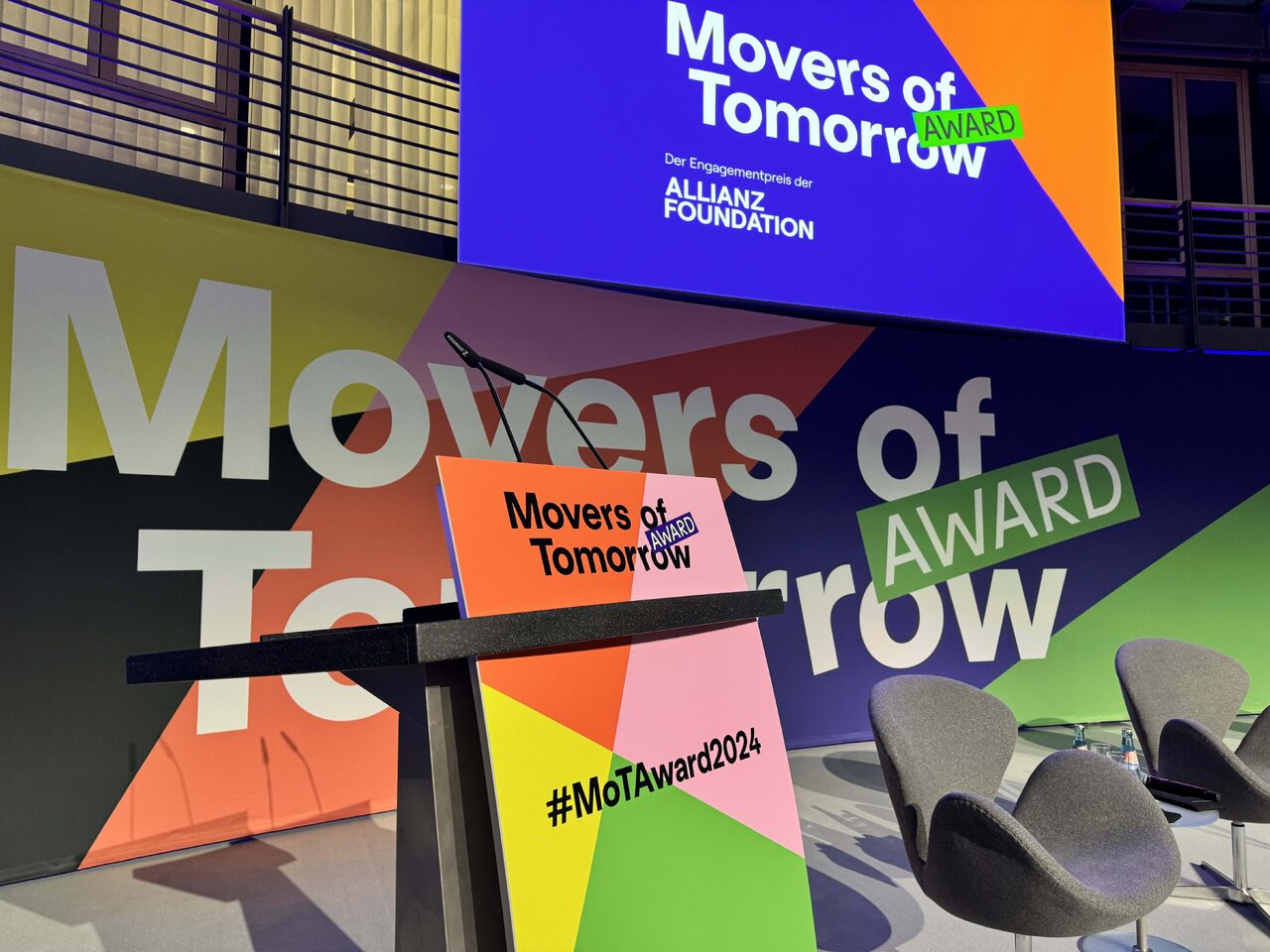 Grafische Darstellung des Movers of Tomorrow Awards mit mehrfarbigen Oberfl&auml;chen und dem Award-Logo in Blau, Orange und Gr&uuml;n, inklusive des Hashtags #MoTAward2024 auf einem Messestand oder einer Pr&auml;sentationsfl&auml;che.