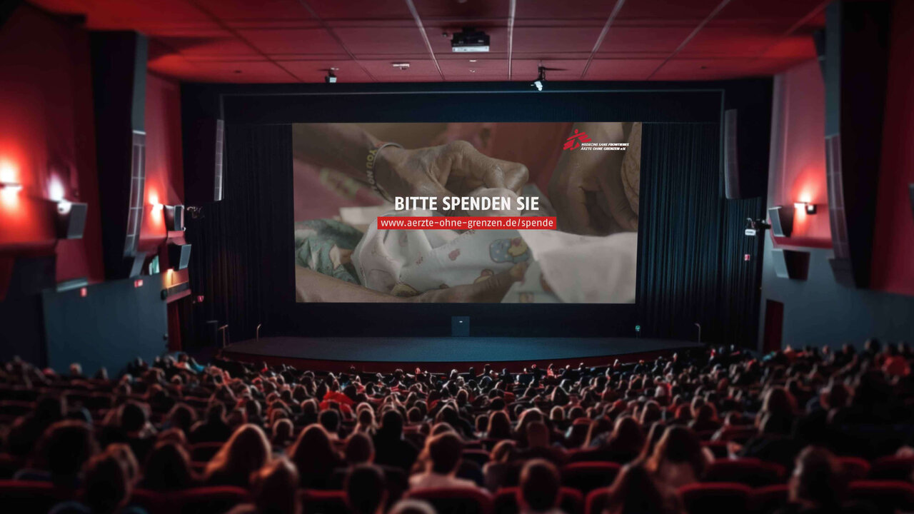 Gro&szlig;er Kinosaal mit vollbesetzten Sitzreihen und einer gro&szlig;en Leinwand, auf der ein Spendenaufruf f&uuml;r eine Hilfsorganisation mit eindringlichem Bild zu sehen ist.