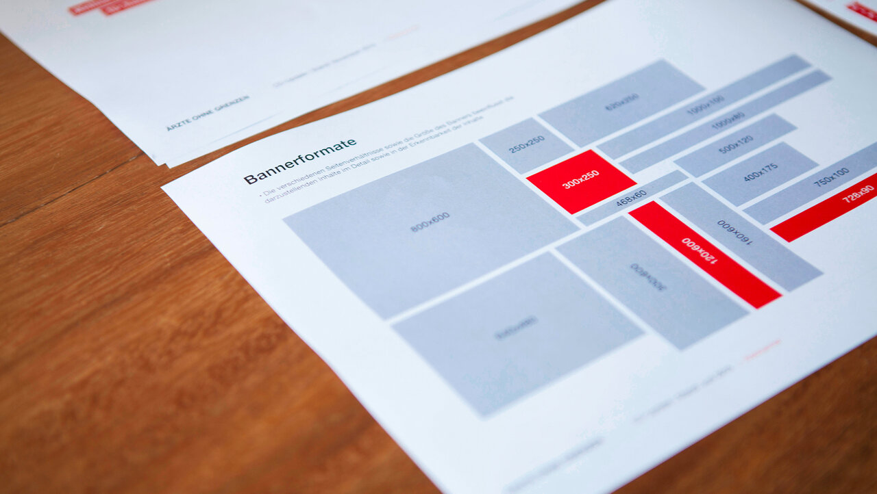 Layout-Skizze eines Webdesigns mit grauen und roten Bl&ouml;cken auf einem Papierdokument, das auf einer Holzoberfl&auml;che liegt. Verschiedene Sektionen und Elemente des Wireframes sind in unterschiedlichen Graut&ouml;nen und markanten Rot-Akzenten gestaltet.