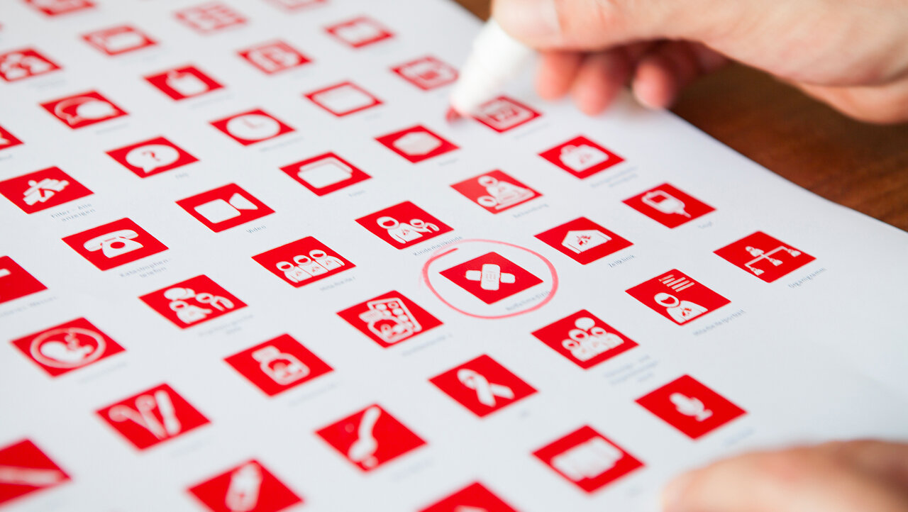 Rote und wei&szlig;e Raster mit Icons, die verschiedene Symbole und Funktionen darstellen, w&auml;hrend eine Hand mit einem Stift ein Element auf dem Blatt ausw&auml;hlt oder markiert.
