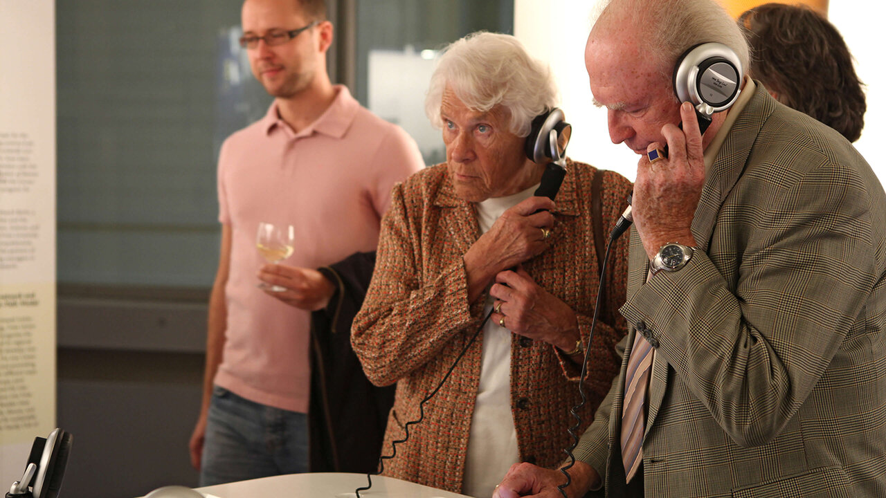 Zwei &auml;ltere Menschen mit Kopfh&ouml;rern und einem j&uuml;ngeren Mann im Hintergrund, die sich konzentriert an einem Ausstellungsstand mit technischen Ger&auml;ten befinden.