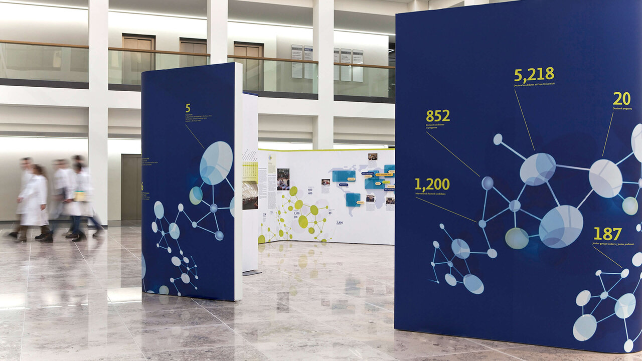 Drei blaue Ausstellungsw&auml;nde mit vernetzten Knotenpunkten und Zahlen, die eine wissenschaftliche oder netzwerkartige Struktur darstellen. Im Hintergrund sind verschwommene Personen in wei&szlig;er Kleidung in einem modernen, hellen Ausstellungsraum zu sehen.