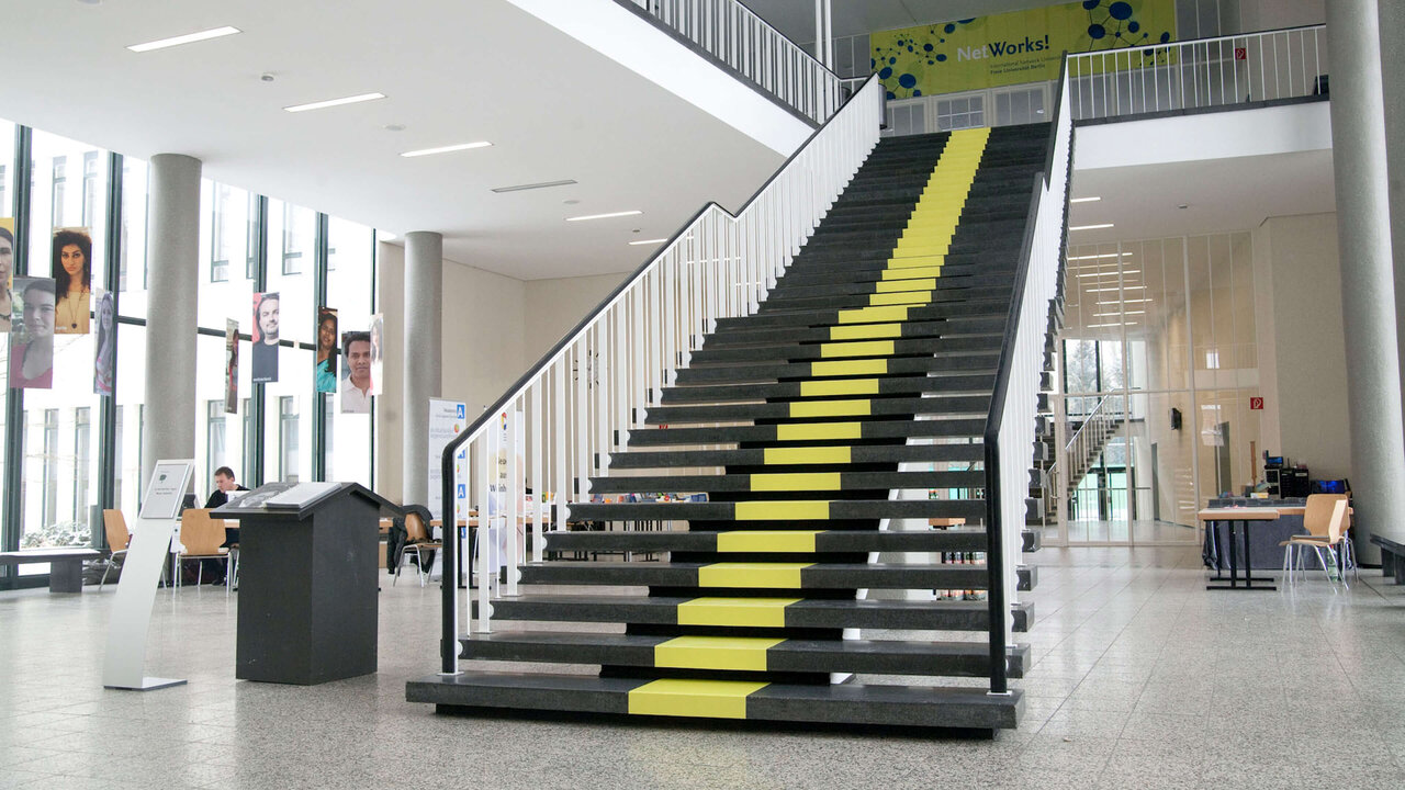 Breite Treppe mit schwarzen und gelben Stufen in einem hellen, modernen Geb&auml;udeeingang. Die Treppe f&uuml;hrt &uuml;ber zwei Etagen und ist Teil eines lichtdurchfluteten Foyers mit Glasfassaden und Mobiliar.