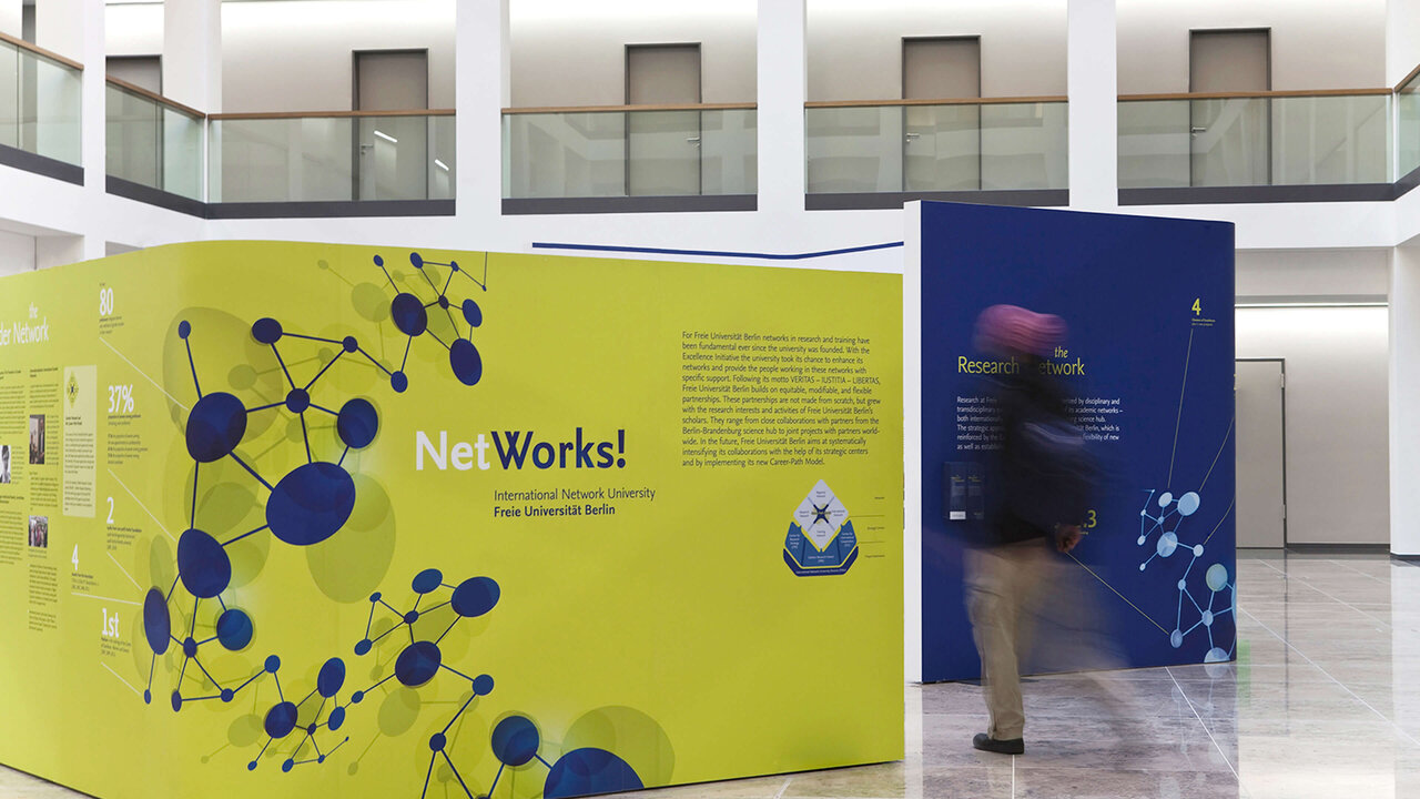 Wanderausstellung &bdquo;NetWorks!" mit grafischer Darstellung von Netzwerkknoten in Gelb und Blau, aufgestellt in einem modernen Ausstellungsraum mit Glasarchitektur. Eine verschwommene Person bewegt sich an den Ausstellungsw&auml;nden vorbei, die Informationen &uuml;ber die Freie Universit&auml;t Berlin pr&auml;sentieren.