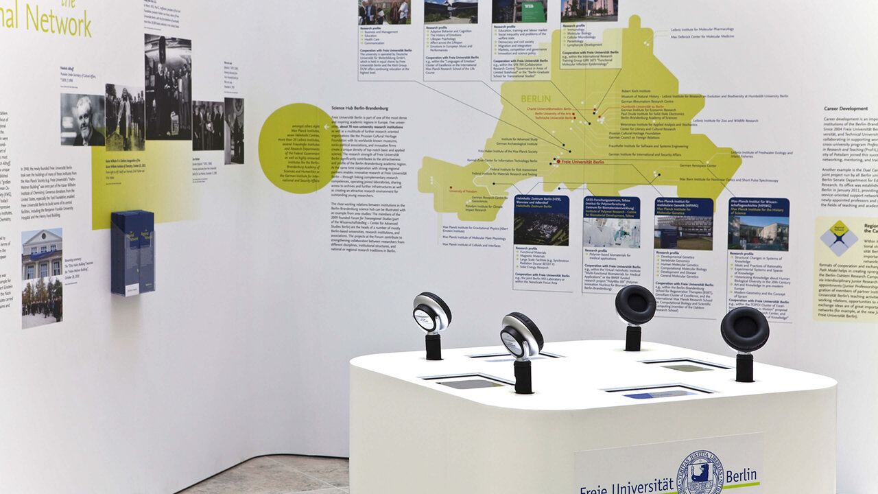 Ausstellungsinstallation mit gelbem Zeitstrahl und wei&szlig;em Ausstellungstisch mit vier schwarzen Mikrofonen, die Netzwerkuniversit&auml;t und Forschungsgeschichte der Freien Universit&auml;t Berlin dokumentierend.