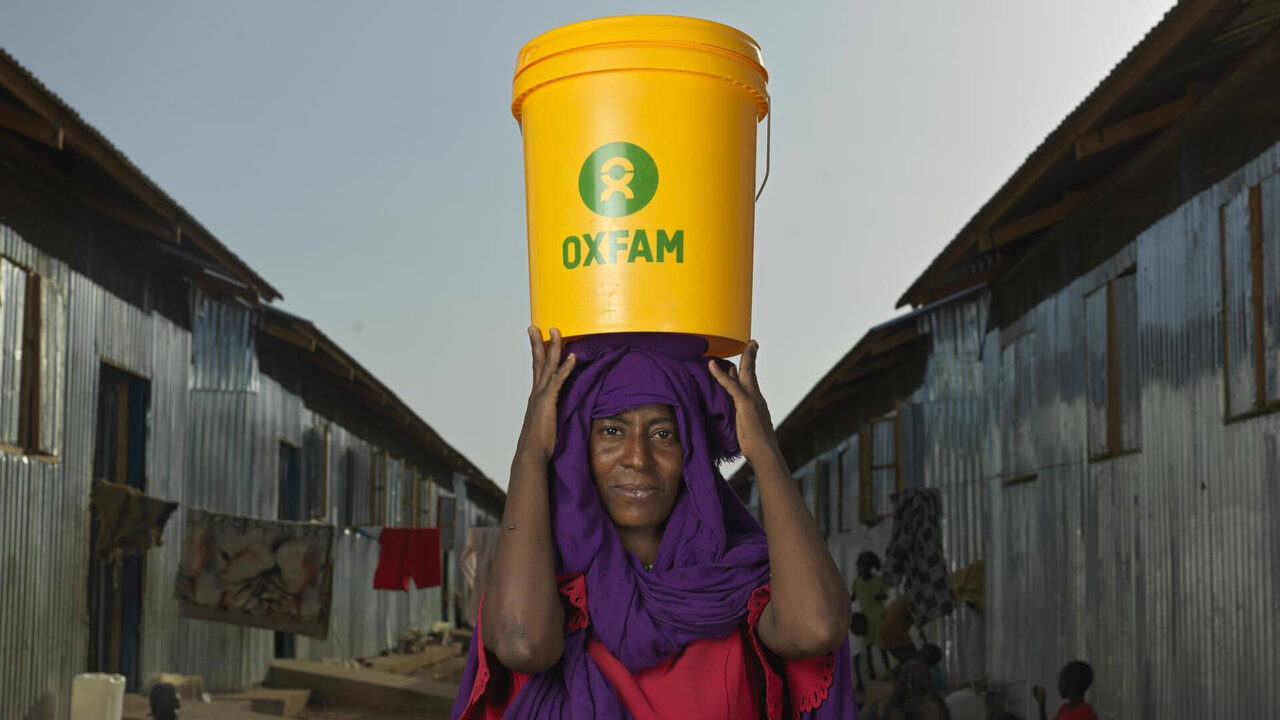 Eine Person mit violettem Schal und roter Kleidung tr&auml;gt einen gelben Oxfam-Eimer auf dem Kopf, umgeben von grauen Wellblechh&uuml;tten in einer scheinbar &auml;rmlichen Umgebung.