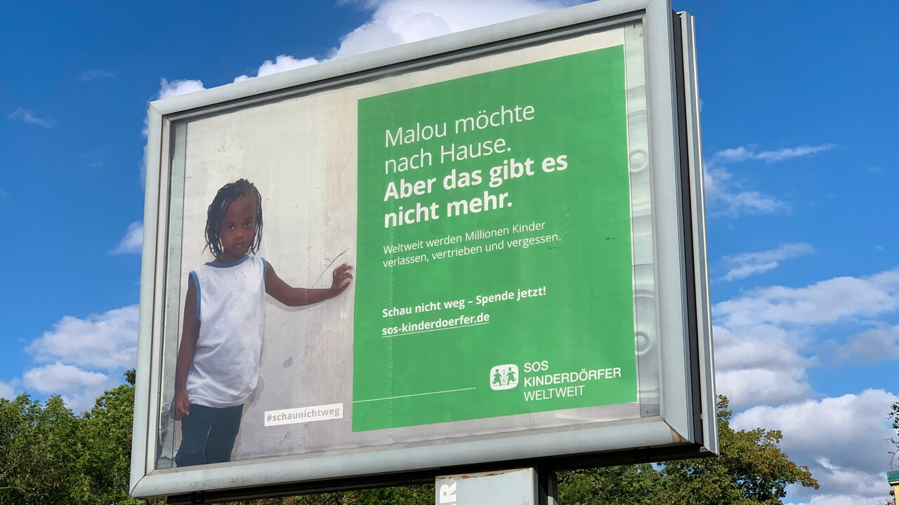 Gro&szlig;formatiges Werbeplakat f&uuml;r SOS-Kinderd&ouml;rfer mit einer Person in wei&szlig;em T-Shirt vor gr&uuml;nem Hintergrund. Der Text "Malou m&ouml;chte nach Hause. Aber das gibt es nicht mehr." steht neben der abgebildeten Person und ruft zum Spenden auf.