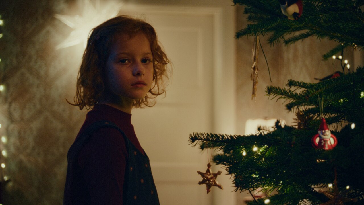Ein junges M&auml;dchen in einem dunklen Pullover steht nachdenklich vor einem Weihnachtsbaum mit Lichtern und Ornamenten, wobei ein ged&auml;mpftes Licht die melancholische Stimmung unterstreicht.