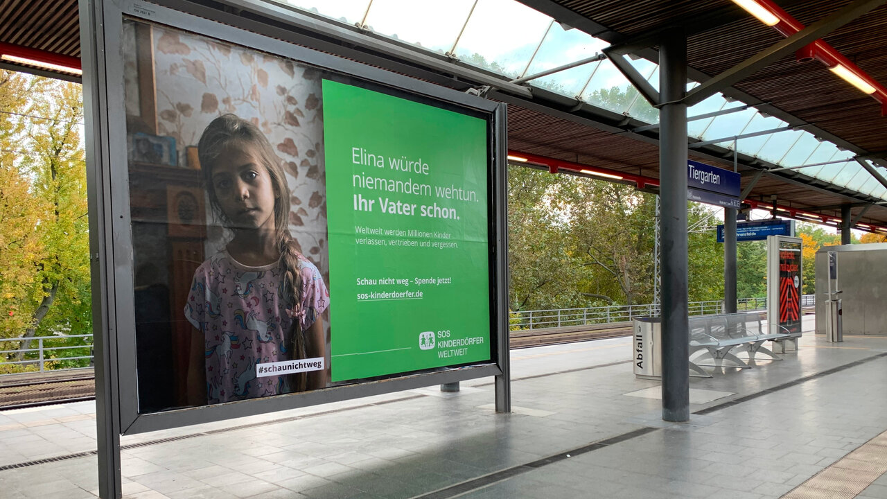Werbeplakat an einer Bahnstation mit einem Foto eines Kindes neben einer gr&uuml;nen Farbfl&auml;che, das auf die Unterst&uuml;tzung von Kindern durch SOS-Kinderd&ouml;rfer aufmerksam macht. Der Slogan suggeriert einen emotionalen Appell zur Hilfe f&uuml;r Kinder in Not.