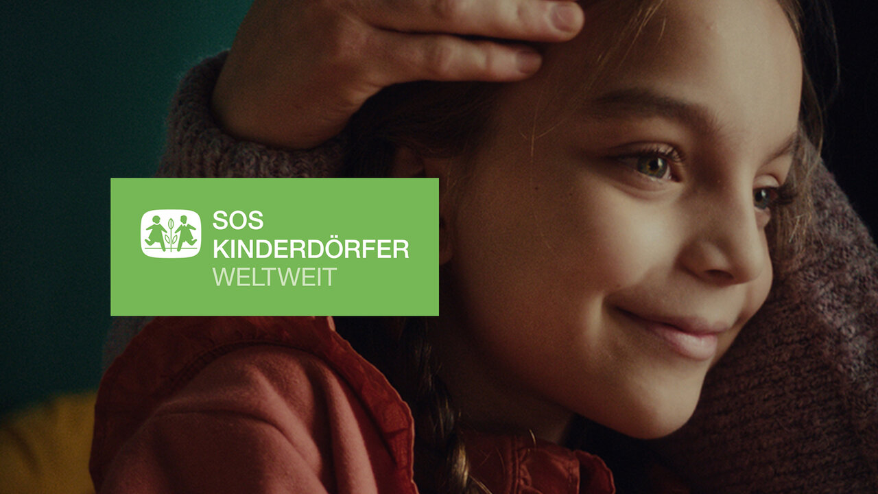 Portr&auml;t eines l&auml;chelnden Kindes in warmen Rott&ouml;nen, das von weichem Licht umgeben ist, mit dem Logo der SOS-Kinderd&ouml;rfer weltweit in gr&uuml;nem Kasten.