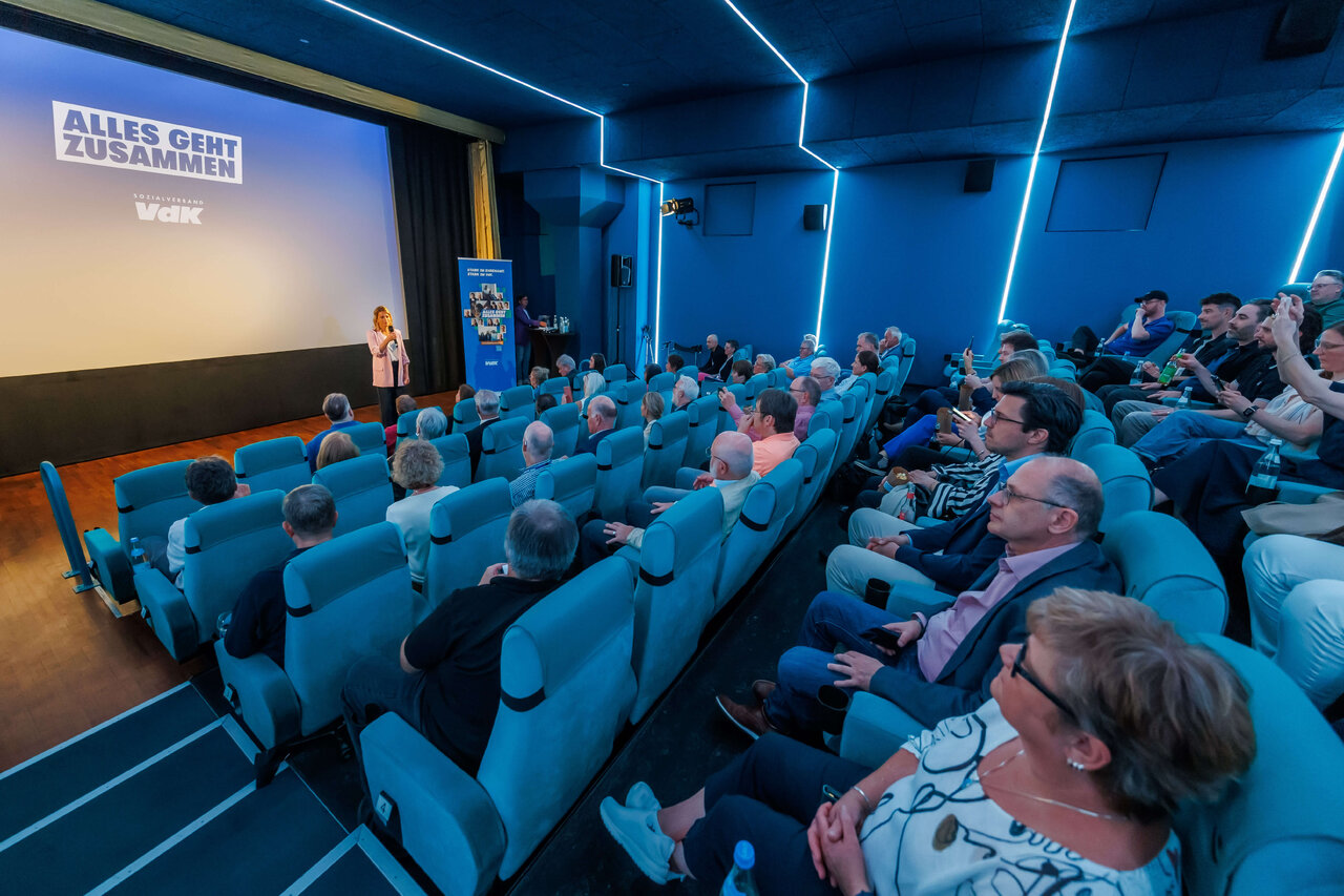 Konferenzraum mit blau beleuchteten W&auml;nden und einem Podium, auf dem eine Person spricht. Publikum sitzt in blauen Sitzen und h&ouml;rt aufmerksam zu, w&auml;hrend auf der Leinwand eine Pr&auml;sentation l&auml;uft.