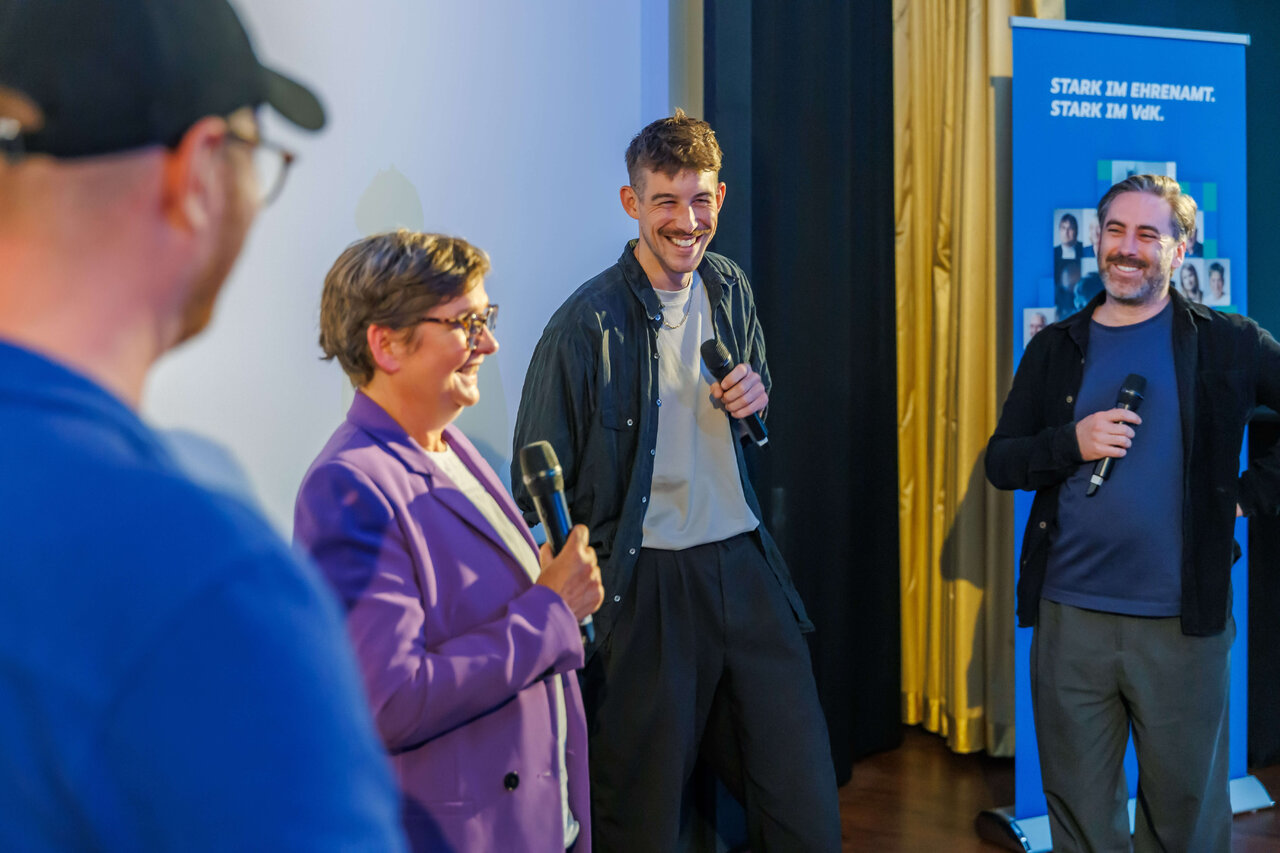 Drei Personen bei einer Pr&auml;sentation oder Diskussionsrunde, eine Frau in einem lila Blazer und zwei M&auml;nner, die Mikrofone halten und l&auml;chelnd vor einem blauen Hintergrund mit Vorh&auml;ngen stehen.