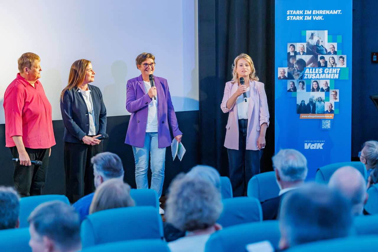 Vier Frauen stehen auf einer B&uuml;hne vor einem blauen Veranstaltungsplakat, mit Publikum in blauen Sitzreihen im Vordergrund. Sie tragen Blazer in verschiedenen Farben und halten Mikrofone w&auml;hrend einer Podiumsdiskussion.