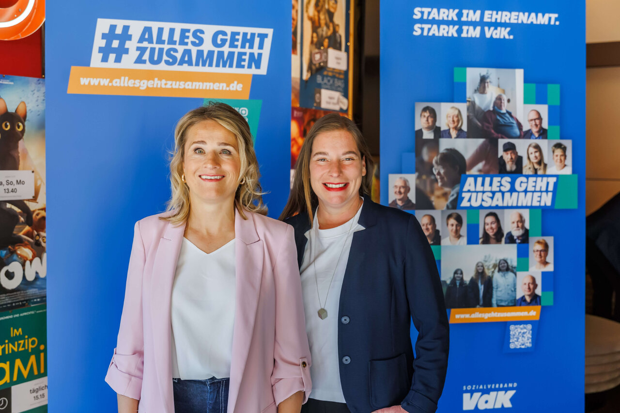 Zwei l&auml;chelnde Frauen in Business-Kleidung vor blauen Plakaten mit dem Slogan "Alles geht zusammen" und Gruppenfotos im Hintergrund, offensichtlich bei einer Veranstaltung oder Kampagne.