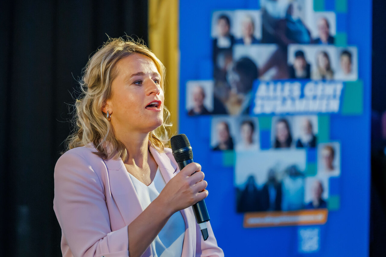 Eine Frau in einem rosa Blazer spricht in ein Mikrofon vor einem blauen Hintergrund mit Portraitfotos. Sie scheint eine Pr&auml;sentation oder Rede bei einer Veranstaltung zu halten.