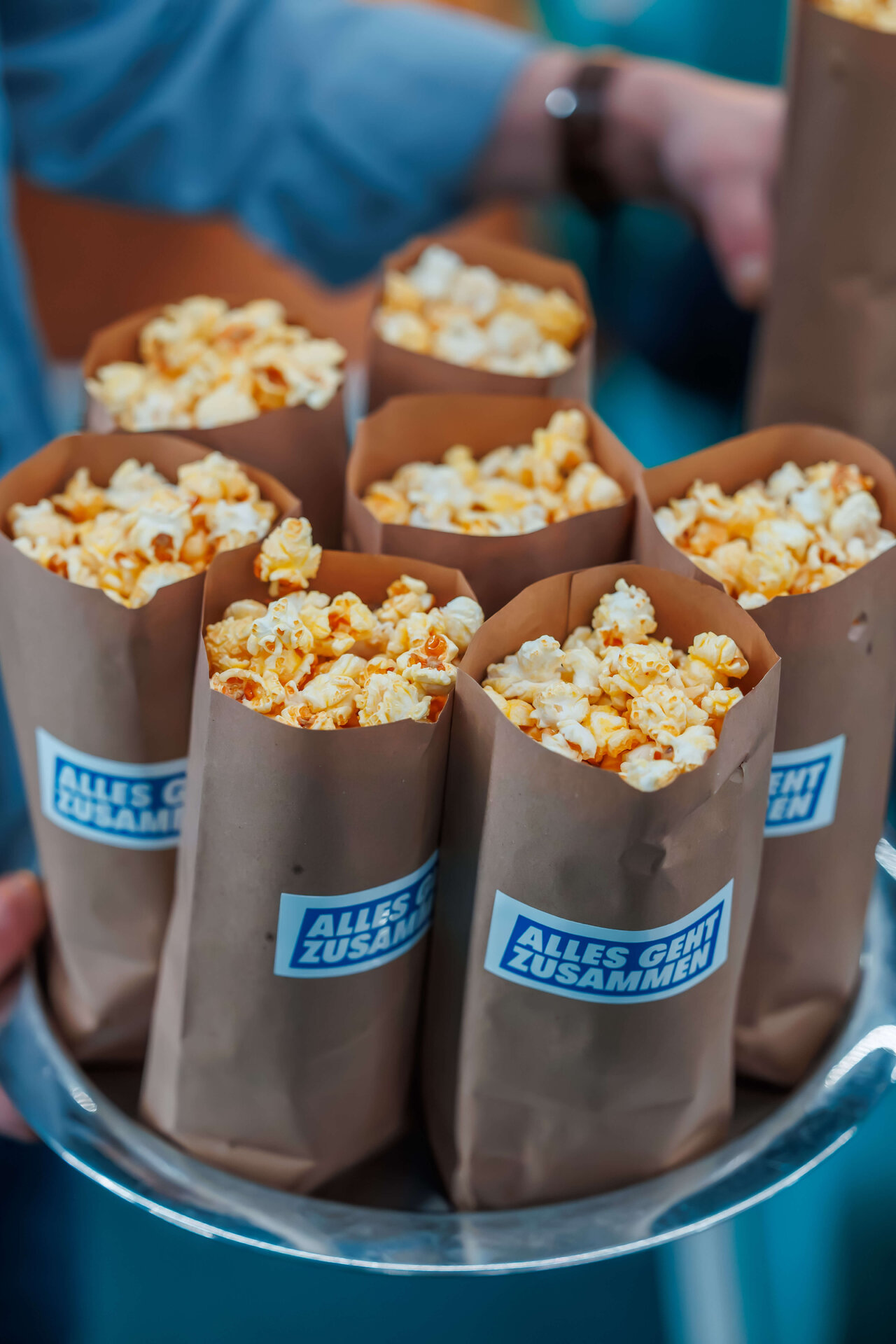 Sieben braune Papiert&uuml;ten gef&uuml;llt mit gelblich-wei&szlig;em Popcorn, die mit einem blauen "Alles gut zusammen" Aufkleber versehen sind, auf einem runden Tablett angeordnet.