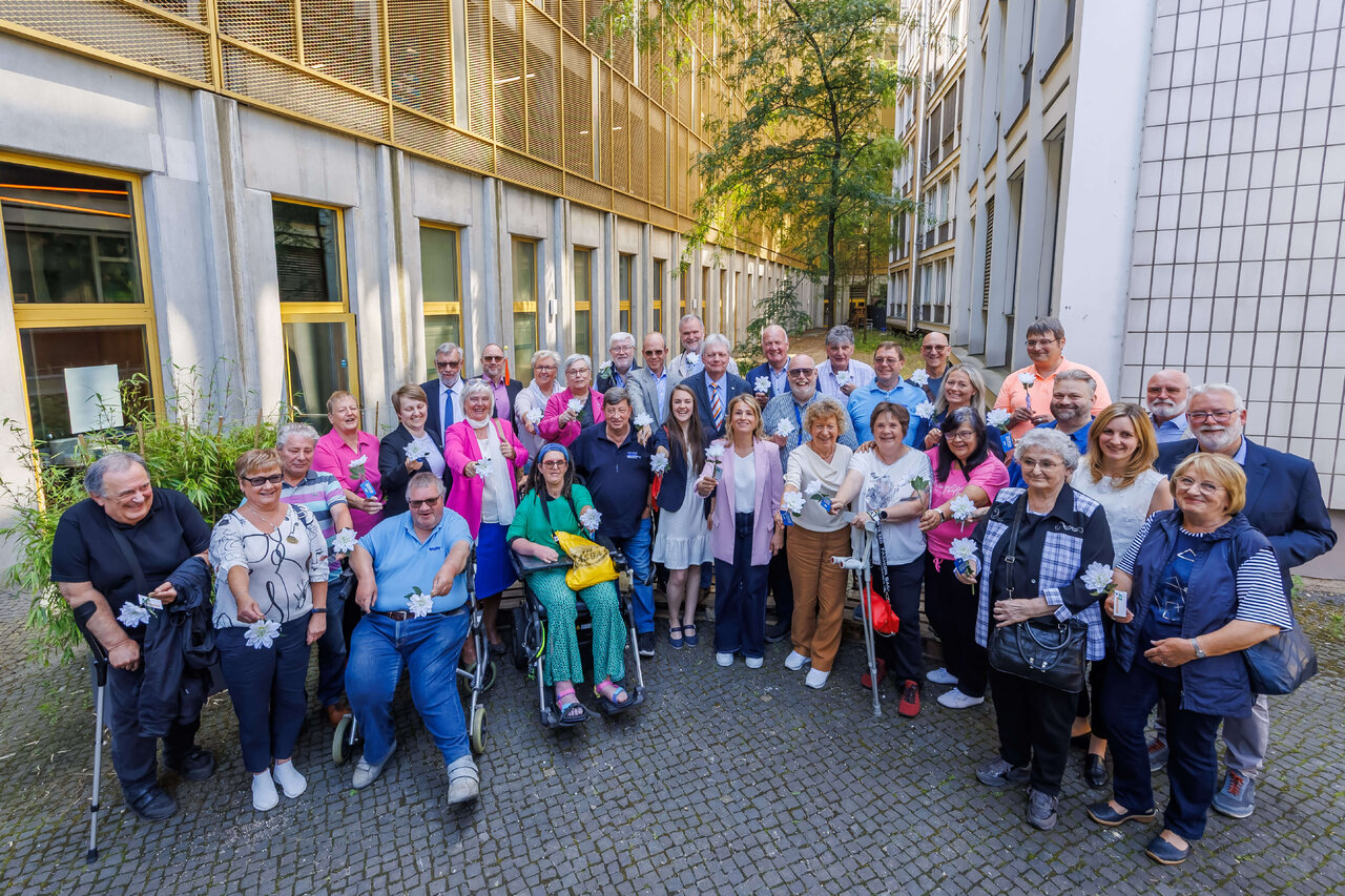 Gro&szlig;e Gruppe von Menschen unterschiedlichen Alters und mit verschiedenen F&auml;higkeiten stehen gemeinsam vor einem Geb&auml;ude, viele tragen bunte Kleidung und einige sitzen im Rollstuhl. Die Gruppe wirkt vielf&auml;ltig, inklusiv und verbunden, mit einem Mix aus Generationen und Hintergr&uuml;nden. Sie repr&auml;sentieren Zusammenhalt und Gemeinschaft in einem urbanen Umfeld.