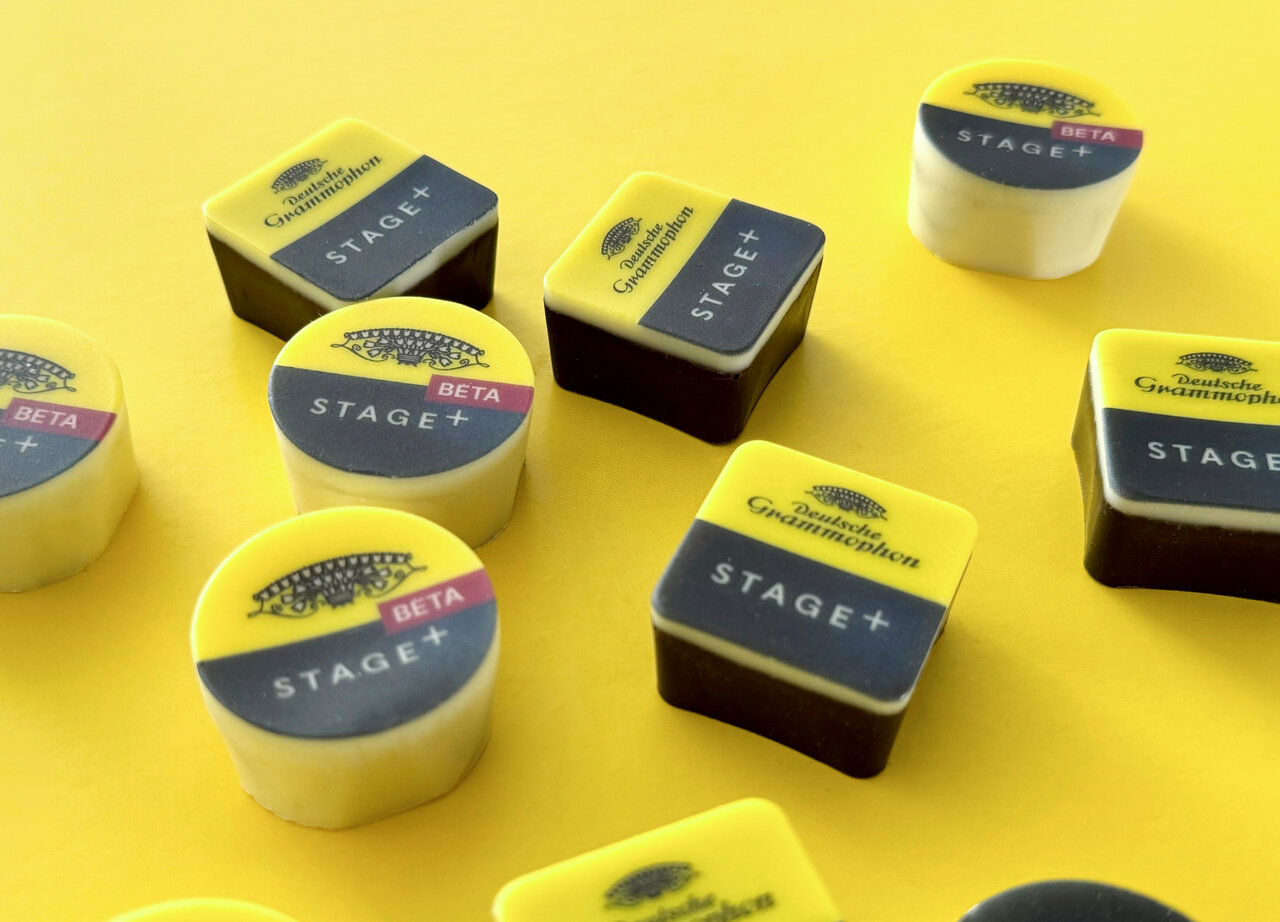 Mehrere quadratische und runde Pralinen in Gelb und Schwarz mit dem Logo "Stage+" auf gelbem Untergrund, die unterschiedlich angeordnet und gestapelt sind.