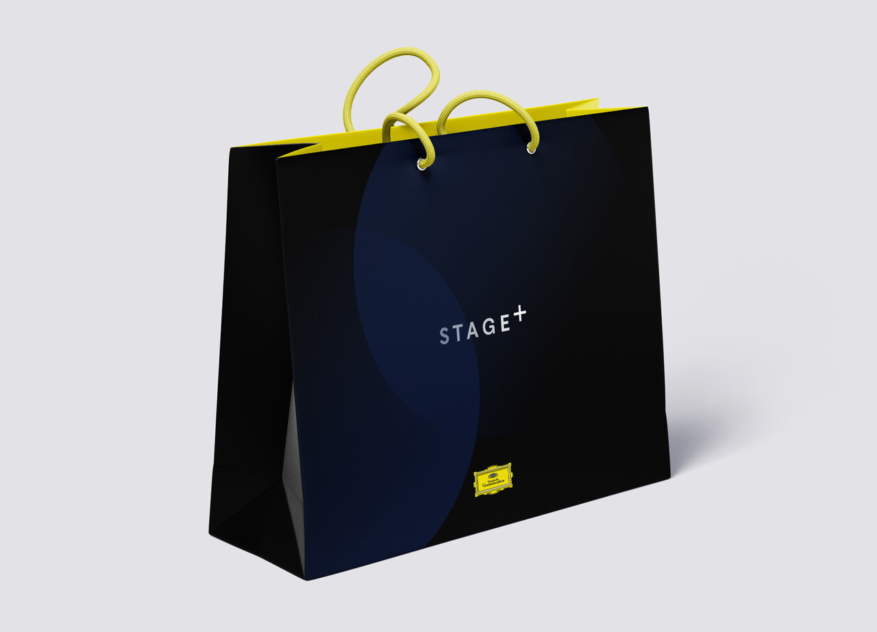 Schwarze Einkaufstasche der Marke Stage+ mit gelben Griffen und einem kleinen gelben Logo, vor einem neutralen Hintergrund pr&auml;sentiert.