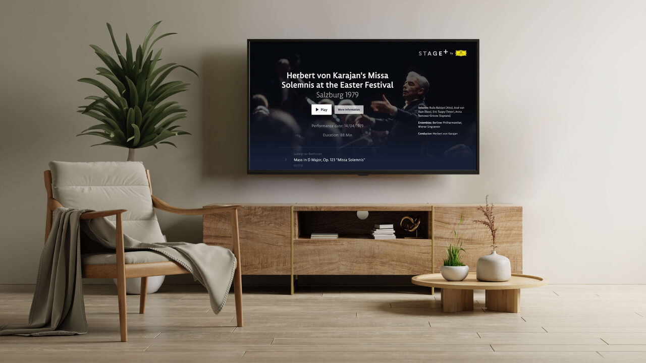 Modernes Wohnzimmer mit einem h&ouml;lzernen Sideboard, einem grauen Sessel und einem Smart-TV, der einen Streaming-Dienst f&uuml;r klassische Musik anzeigt, vor einer neutralen Wand mit einer gro&szlig;en Zimmerpflanze.