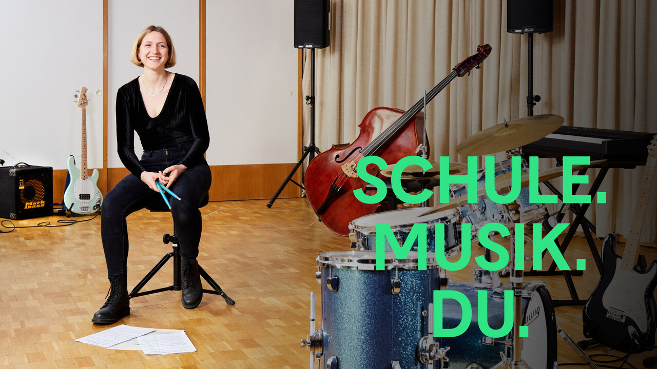 Eine Frau sitzt auf einem Stuhl in einem Musikstudio mit Gitarren, Schlagzeug und dem gr&uuml;nen Schriftzug "SCHULE. MUSIK. DU." im Hintergrund, der die Kampagne f&uuml;r Musiklehrer-Studieng&auml;nge an der Universit&auml;t der K&uuml;nste Berlin symbolisiert.