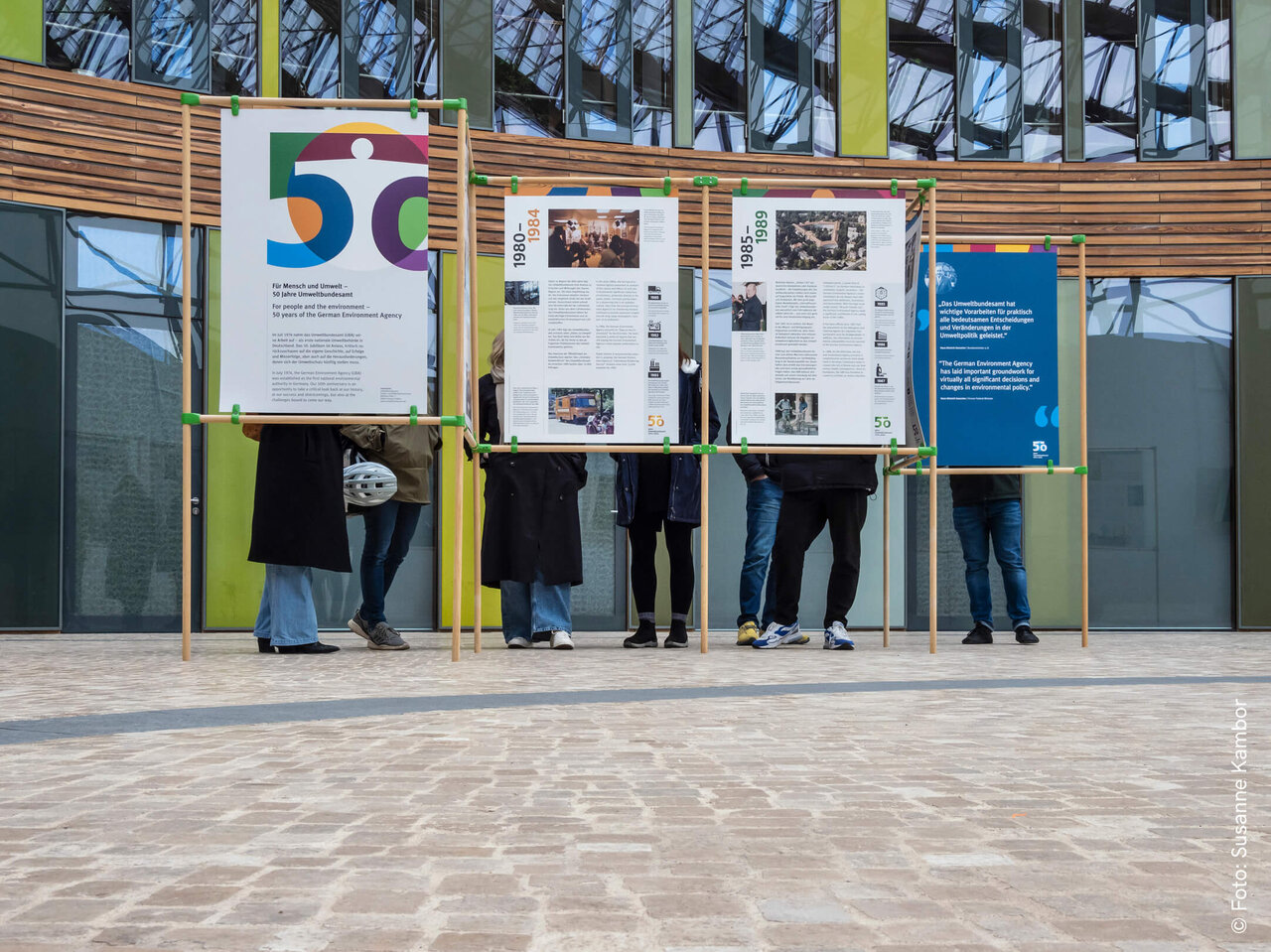 Vier Personen stehen vor Informationstafeln mit verschiedenen Texten und Grafiken, die Teil einer Ausstellung zu scheinen. Die Tafeln sind in Holzrahmen montiert und befinden sich vor einem modernen Glasgeb&auml;ude mit Holzelementen.