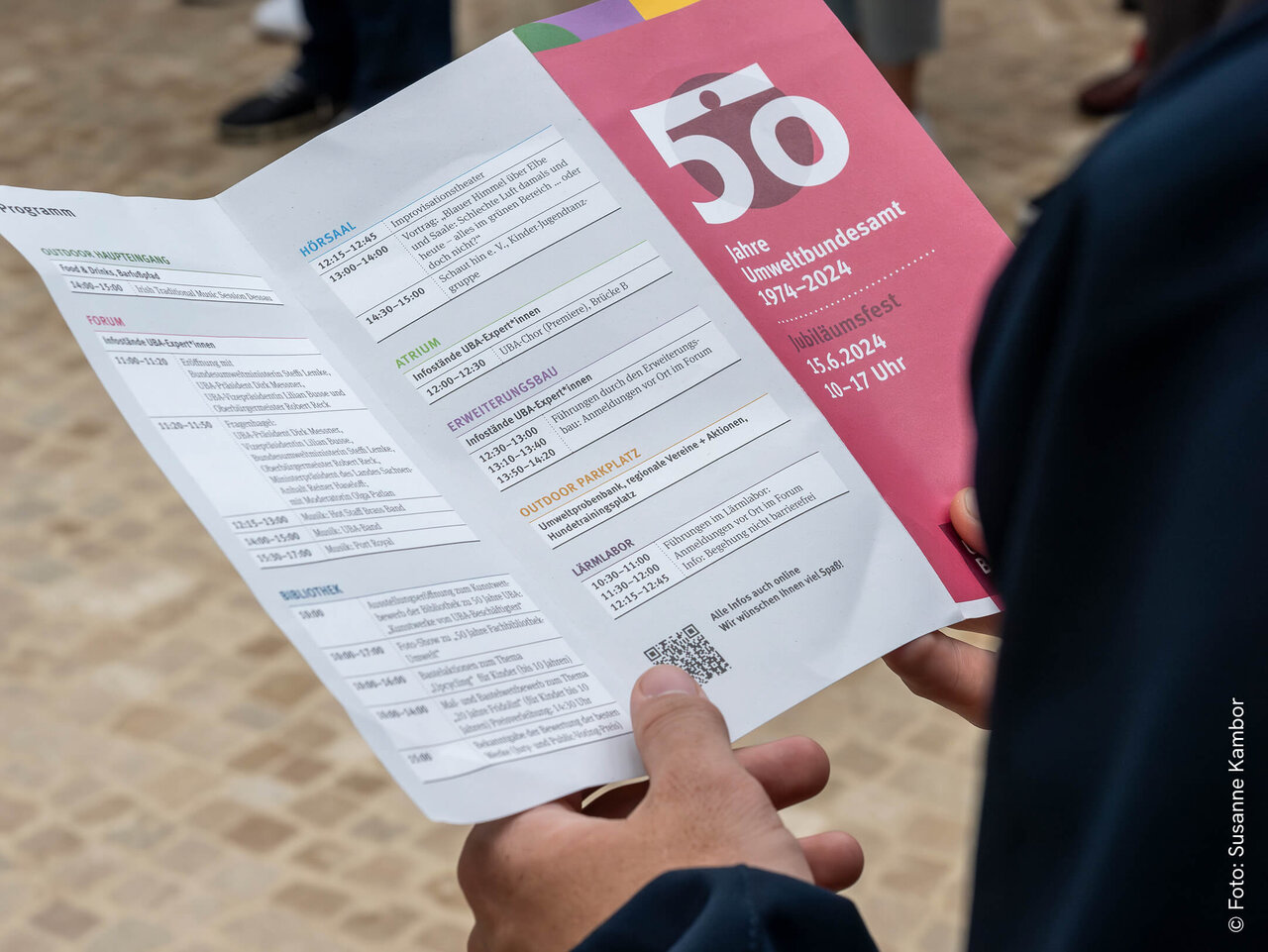 Gefaltetes Informationsfaltblatt mit der Nummer 50 in rosa auf wei&szlig;em Hintergrund, gehalten von einer Hand vor einem unscharfen Hintergrund.