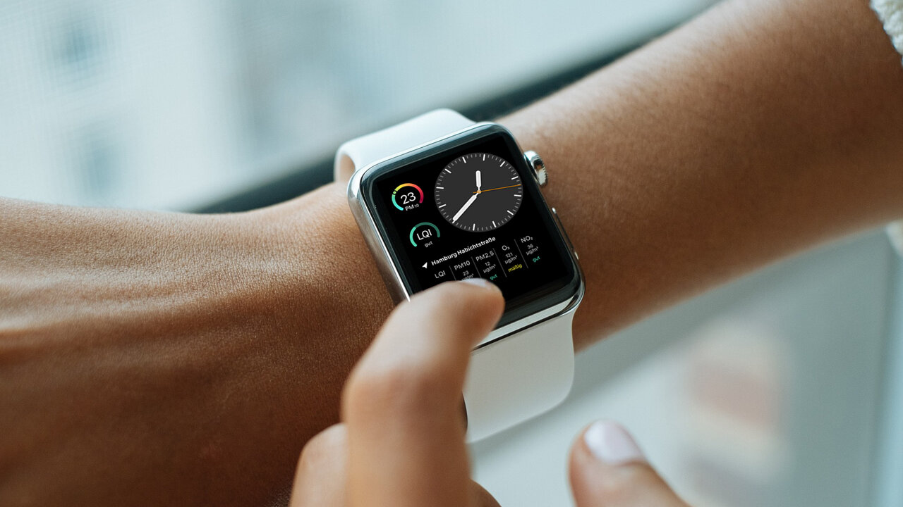 Eine wei&szlig;e Apple Watch mit aktiviertem Bildschirm, die von einer Hand am Handgelenk getragen wird, vor einem unscharfen, hellen Hintergrund.