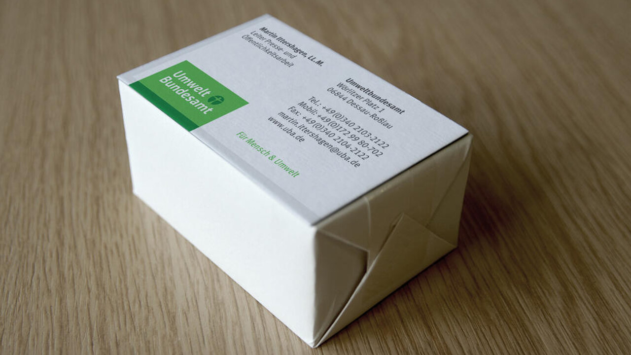 Wei&szlig;e Kartonschachtel mit gr&uuml;nem Etikett auf einer Holzoberfl&auml;che, die eine Produktverpackung oder Publikation von Studio Good f&uuml;r das Umweltbundesamt darstellt.