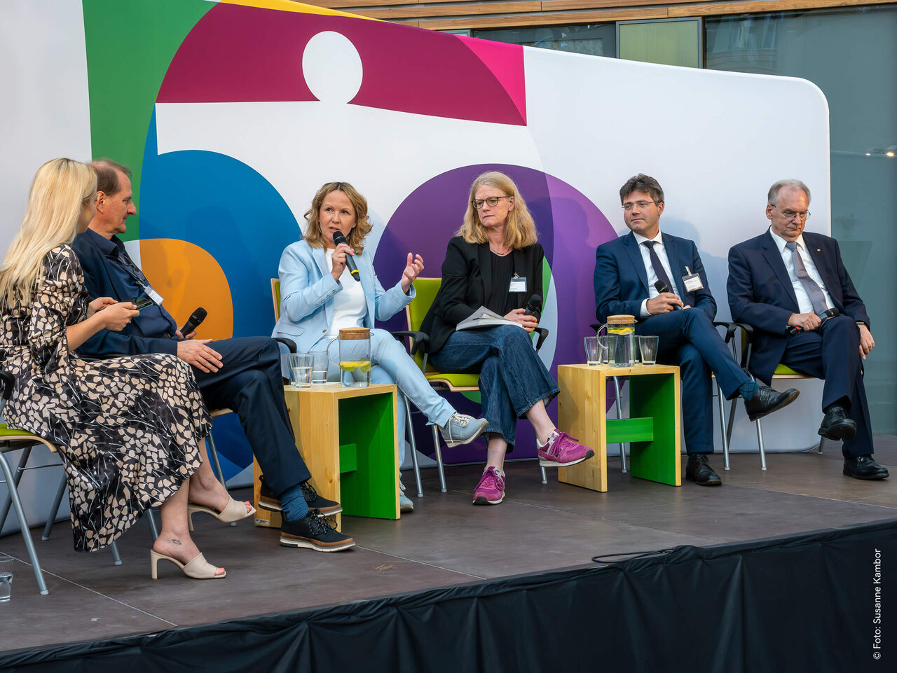 F&uuml;nf Personen sitzen auf einer B&uuml;hne bei einem Podiumsgespr&auml;ch, vor einem bunten geometrischen Hintergrund. Sie tragen Businesskleidung und scheinen an einer Diskussionsveranstaltung teilzunehmen.