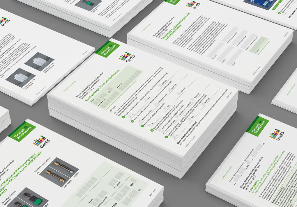 Gestapelte Dokumentenseiten mit gr&uuml;n-wei&szlig;em Layout, die Entw&uuml;rfe und Druckerzeugnisse f&uuml;r das Umweltbundesamt zeigen. Die Bl&auml;tter sind in verschiedenen Gr&ouml;&szlig;en und Ausrichtungen auf einem grauen Hintergrund angeordnet, mit sichtbaren Gestaltungselementen und Textpassagen.