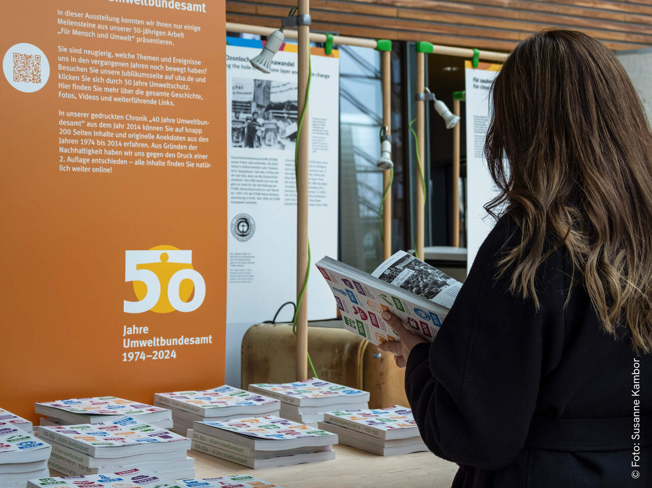 Eine Frau mit langen Haaren steht an einem orangefarbenen Ausstellungsstand, der das 50-j&auml;hrige Jubil&auml;um des Umweltbundesamts markiert. Sie betrachtet ein Informationsmaterial vor Stapeln von Brosch&uuml;ren und Dokumenten.