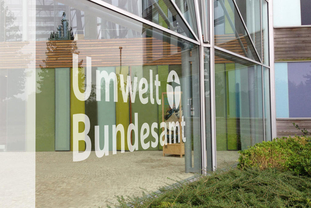 Beschriftung des Umweltbundesamts in gr&uuml;n-gelben Buchstaben an einer modernen Glasfassade mit Holzelementen und einer Gr&uuml;nanlage im Vordergrund.