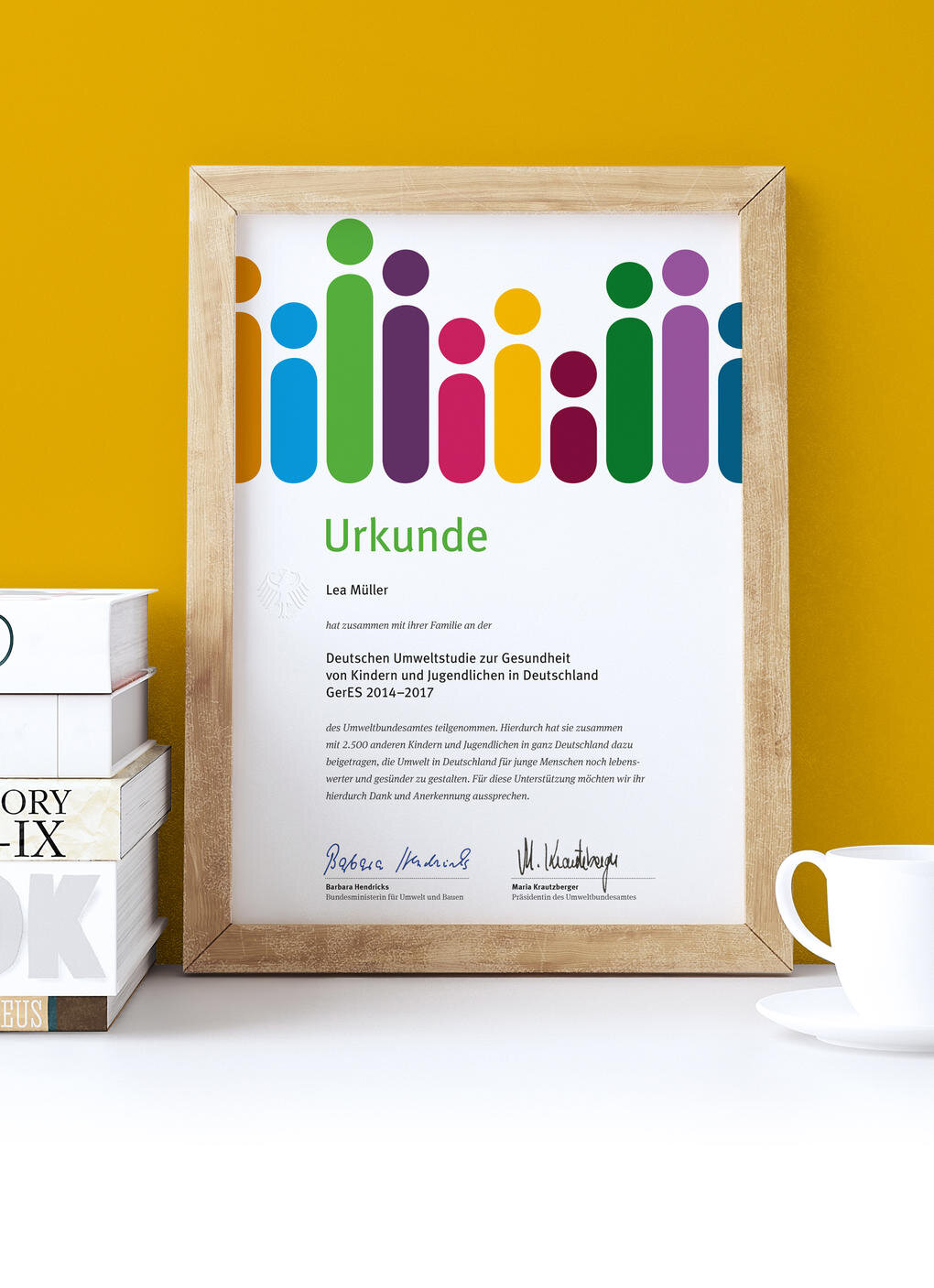 Urkunde des Deutschen Umweltbildungspreises in einem Holzrahmen vor gelbem Hintergrund, mit bunten stilisierten Menschenfiguren als grafisches Element und zwei Unterschriften am unteren Rand.