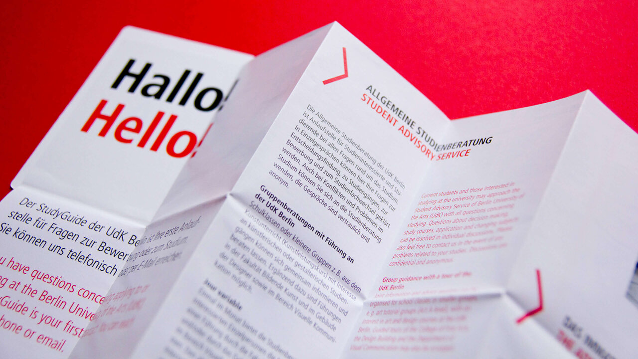 Gefalteter Informationsflyer in Wei&szlig; und Rot mit "Hallo Hello" Schriftzug, der auf einem knallig roten Hintergrund pr&auml;sentiert wird und eine kreative, mehrschichtige Falttechnik zeigt.