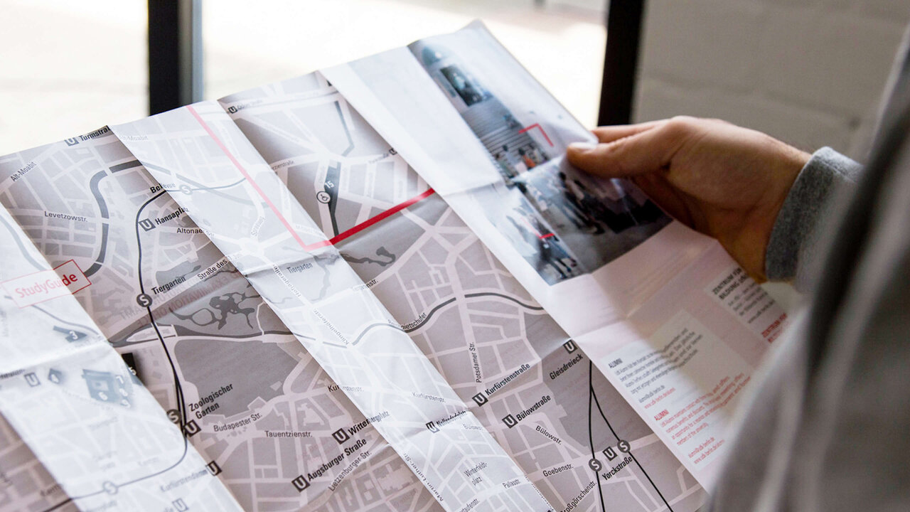 Eine Hand h&auml;lt eine gefaltete Stadtkarte mit Stra&szlig;en und Verkehrswegen, w&auml;hrend der Finger eine bestimmte Route oder Position auf der Karte verfolgt.