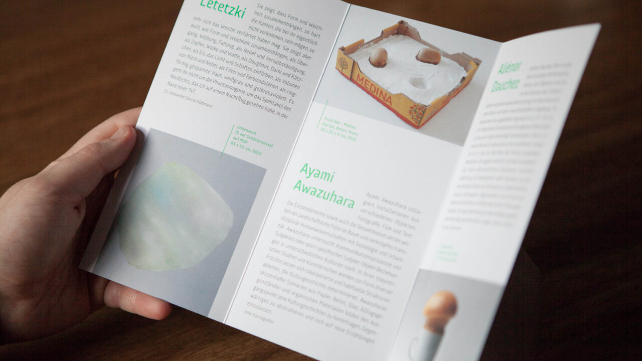 Aufgeschlagene Brosch&uuml;re mit wei&szlig;em Design und gr&uuml;ner Typografie, die einen Text und ein minimalistisches Bild eines Objekts auf einem gefalteten Blatt zeigt, gehalten von einer Hand vor einem braunen Hintergrund.