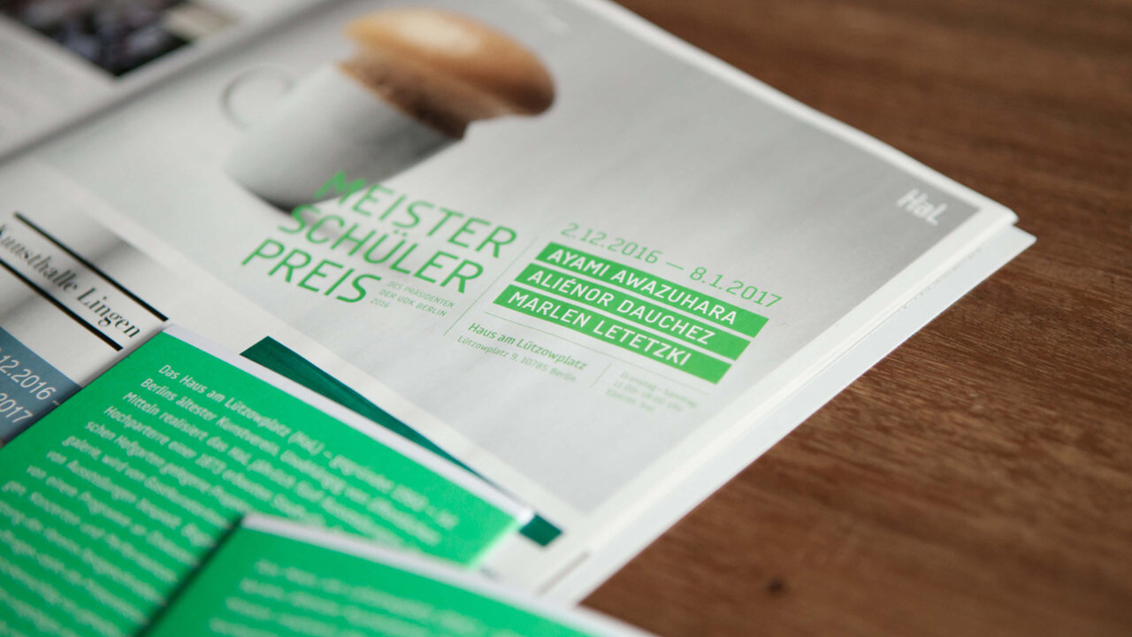 Nahaufnahme eines Druckerzeugnisses mit gr&uuml;ner Typografie, Teil der Gestaltung f&uuml;r den Meistersch&uuml;lerpreis der Universit&auml;t der K&uuml;nste Berlin. Das Dokument liegt auf einer Holzoberfl&auml;che und zeigt einen ausschnittweisen Blick auf das Design.