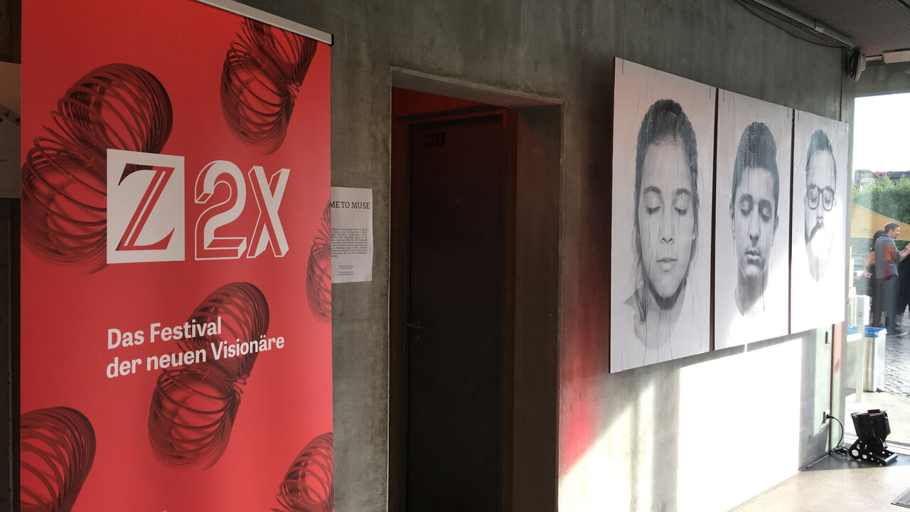 Z2X-Festivalbanner in Rot mit stilisierten Formen und Portr&auml;tfotos an einer Wand, die eine kreative und vision&auml;re Atmosph&auml;re vermitteln.