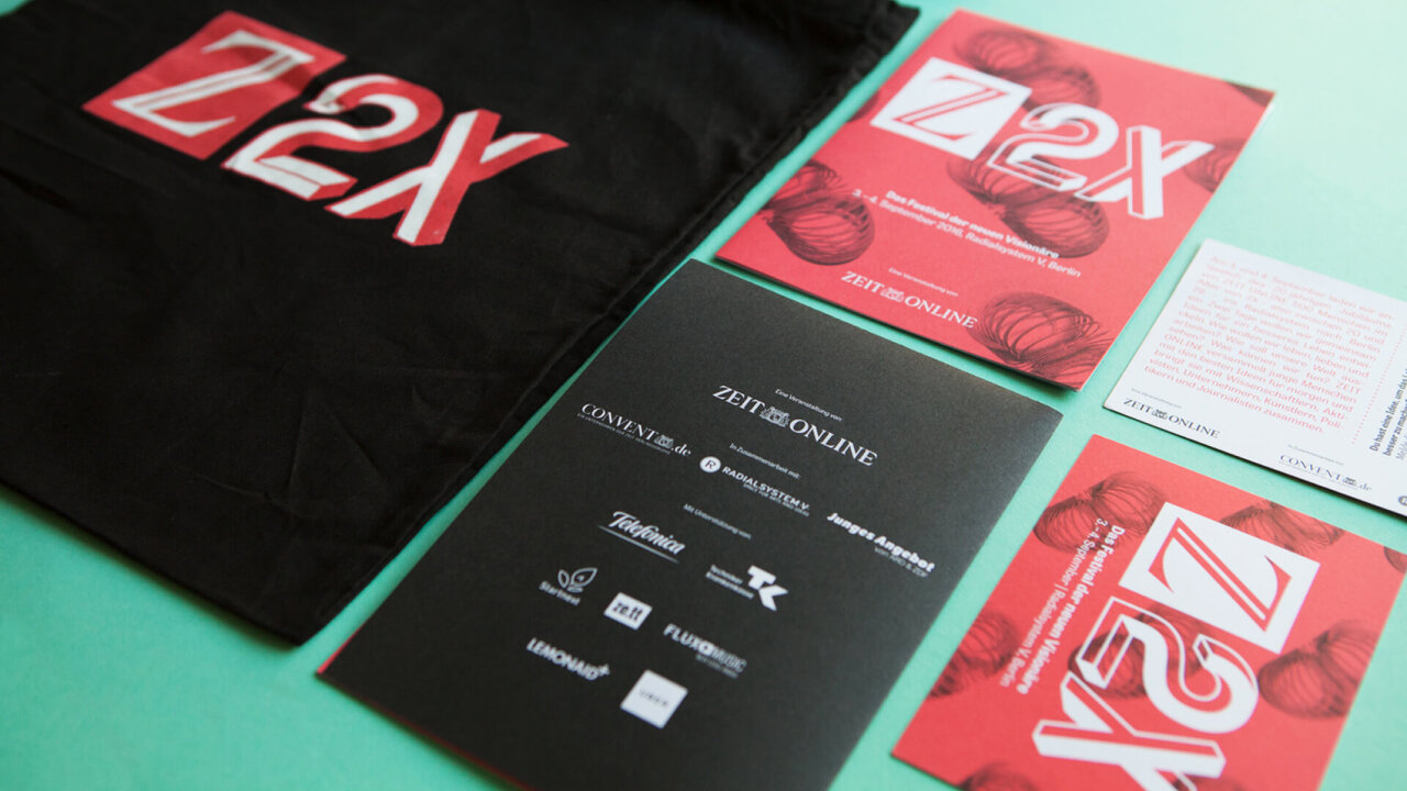 Z2X-Designmaterialien in Rot, Schwarz und T&uuml;rkis, bestehend aus einem schwarzen T-Shirt, Brosch&uuml;ren und einer Informationsseite mit Logos und Veranstaltungsdetails f&uuml;r das Festival der neuen Vision&auml;re von ZEIT ONLINE.