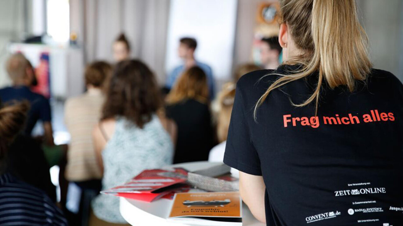 Person von hinten mit schwarzem T-Shirt mit rotem Aufdruck "Frag mich alles", die an einem Tisch in einer Veranstaltung sitzt, umgeben von anderen Teilnehmern im unscharfen Hintergrund.