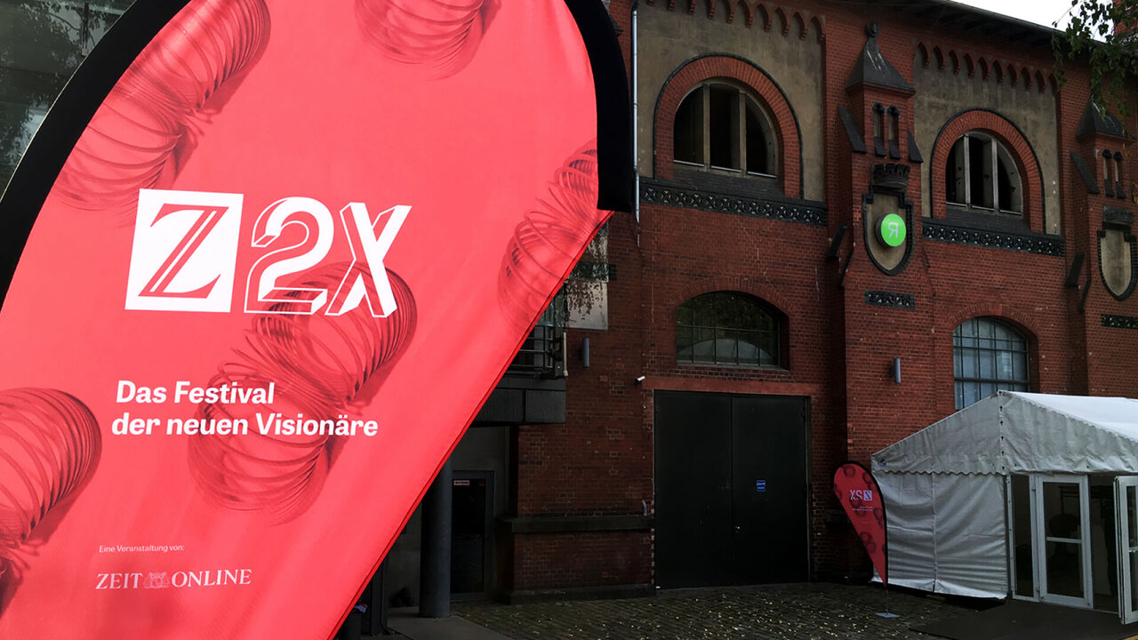 Rote Z2X-Flagge vor einem roten Backsteingeb&auml;ude mit wei&szlig;em Veranstaltungszelt, das das Festival der neuen Vision&auml;re von ZEIT ONLINE ank&uuml;ndigt.