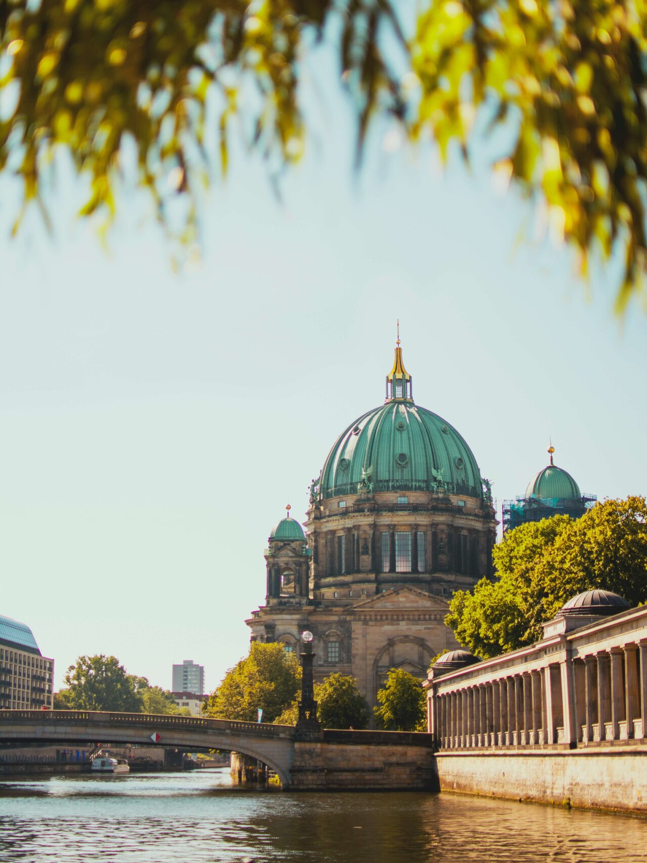 Berliner Dom mit seiner gr&uuml;nen Kuppel, eingebettet in eine sonnendurchflutete Stadtlandschaft mit Spree und B&auml;umen im Vordergrund. Die historische Architektur spiegelt sich im Wasser, w&auml;hrend eine Br&uuml;cke und B&auml;ume die Szenerie rahmen.