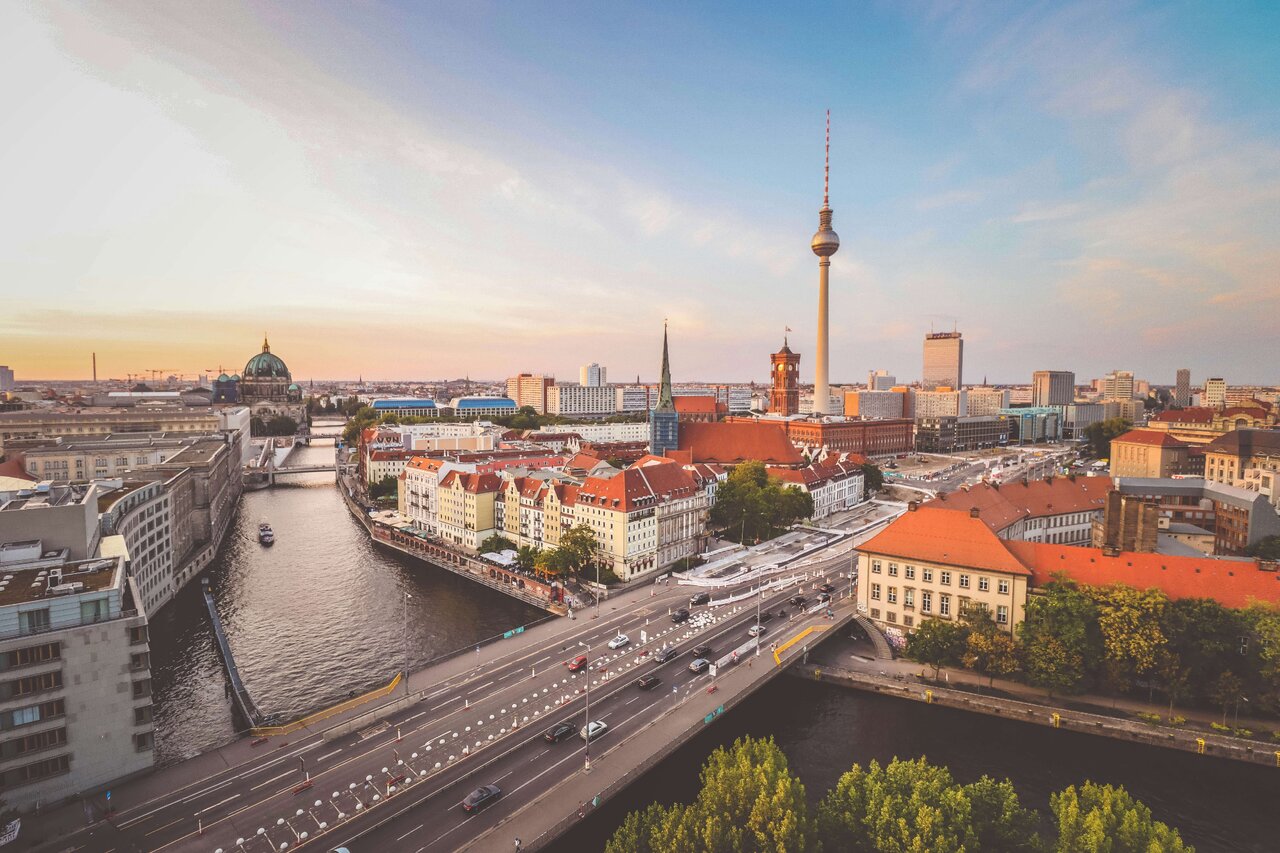 Berliner Stadtpanorama mit Spree, historischen Geb&auml;uden und dem markanten Fernsehturm am Alexanderplatz w&auml;hrend eines goldenen Sonnenuntergangs, der die D&auml;cher und Architektur in warmes Licht taucht.