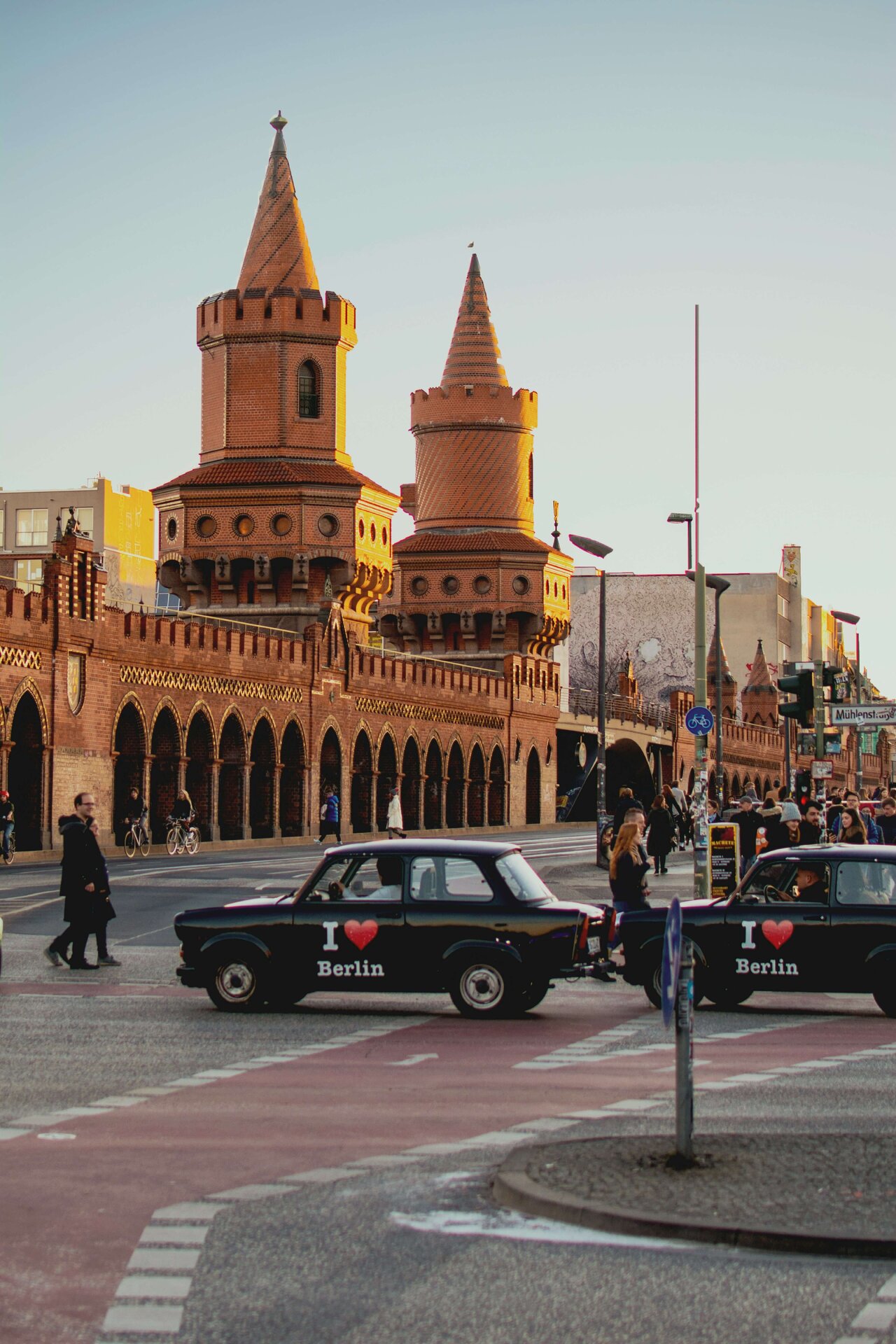 Oberbaumbr&uuml;cke in Berlin mit zwei schwarzen Trabant-Autos mit "I love Berlin" Aufkleber, vor historischer Backsteinarchitektur mit markanten T&uuml;rmen und Arkaden, belebter Stra&szlig;enkreuzung im Vordergrund.
