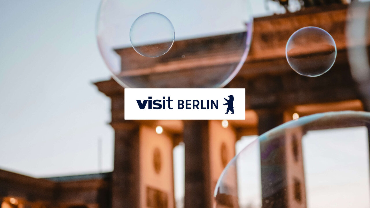 Verschwommene Glasscheiben im Vordergrund mit "visit BERLIN" Logo, dahinter unscharfe Architektur in warmen Braunt&ouml;nen und ged&auml;mpftem Licht.